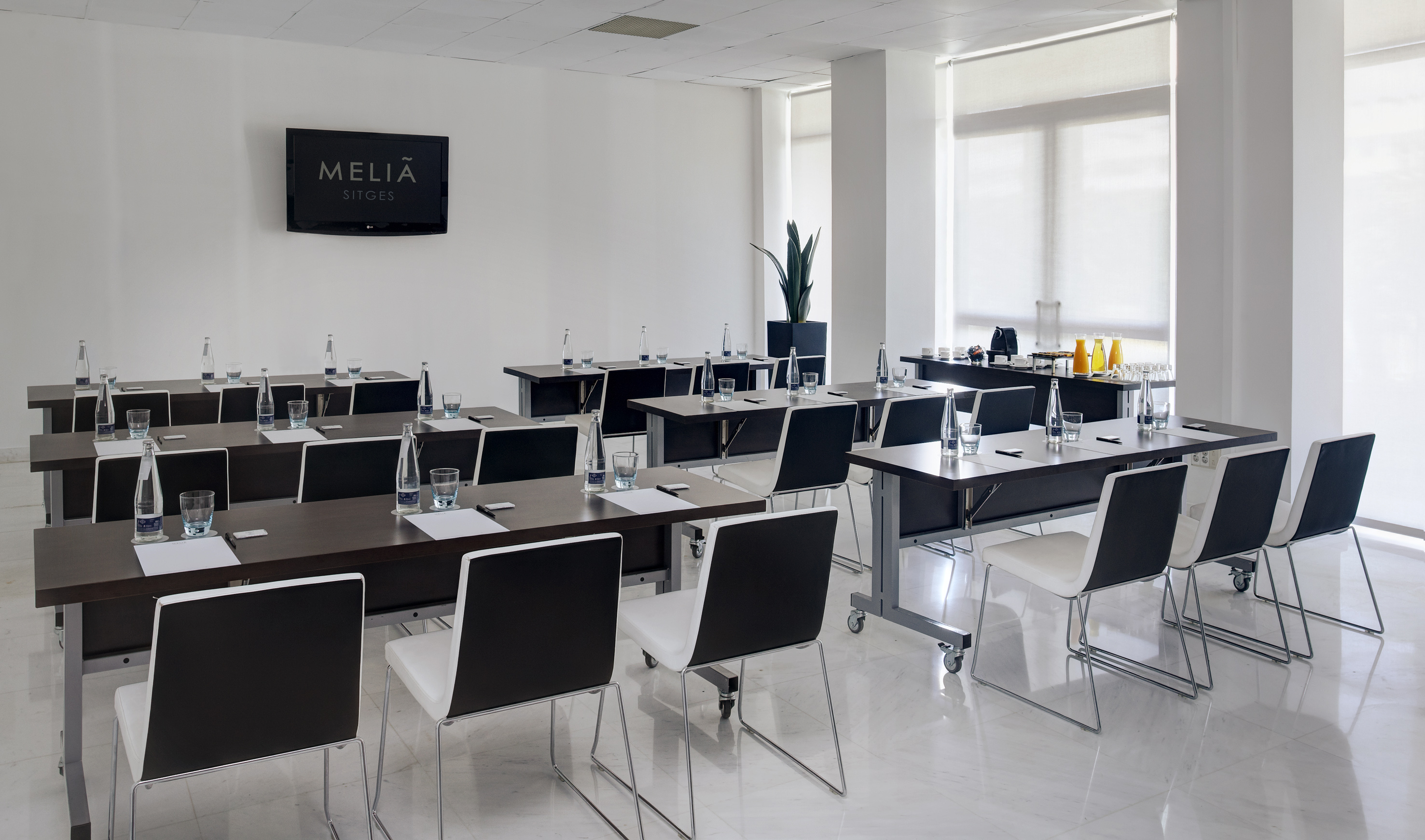 Melia Sitges bright meeting room with classroom setup, TV, and drink station.