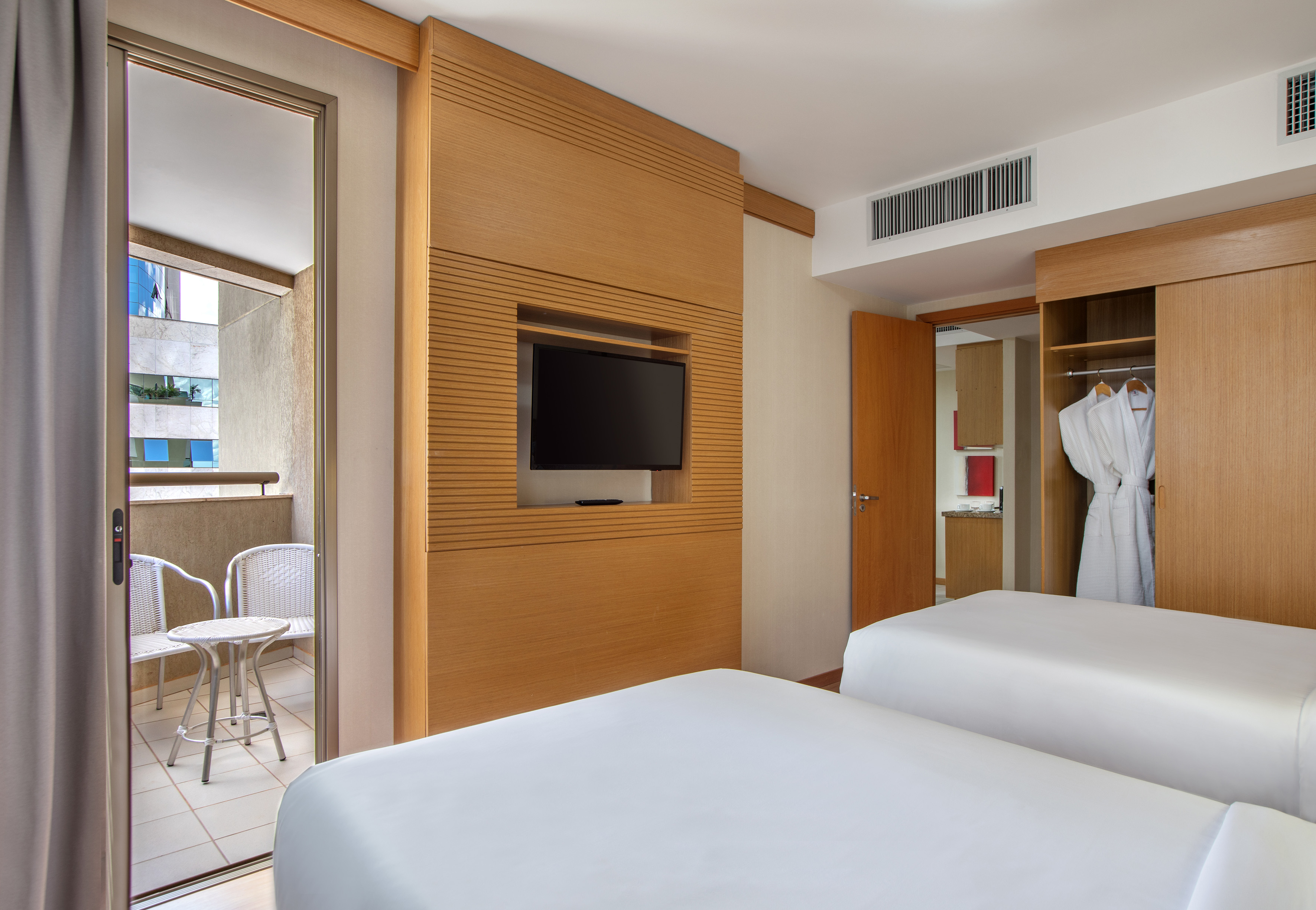 Twin hotel room with wall-mounted TV, open closet, and balcony seating.