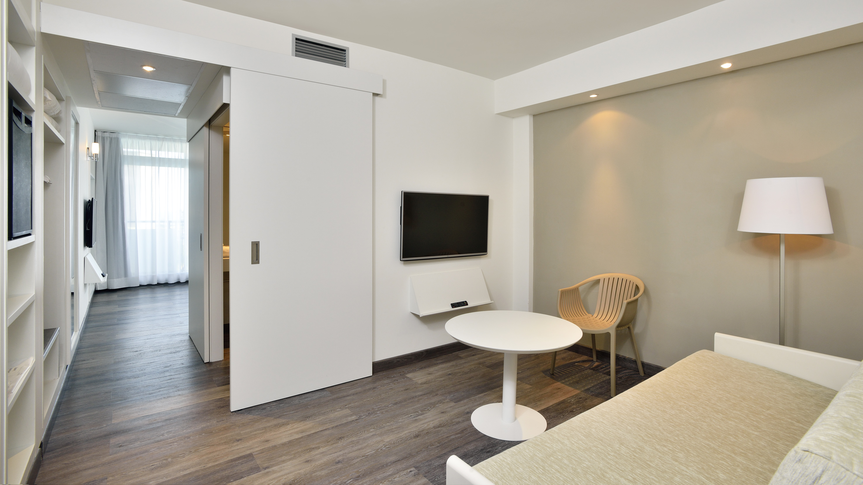Modern hotel room: wall TV, sliding door, table, chair, sofa bed, wood floor.