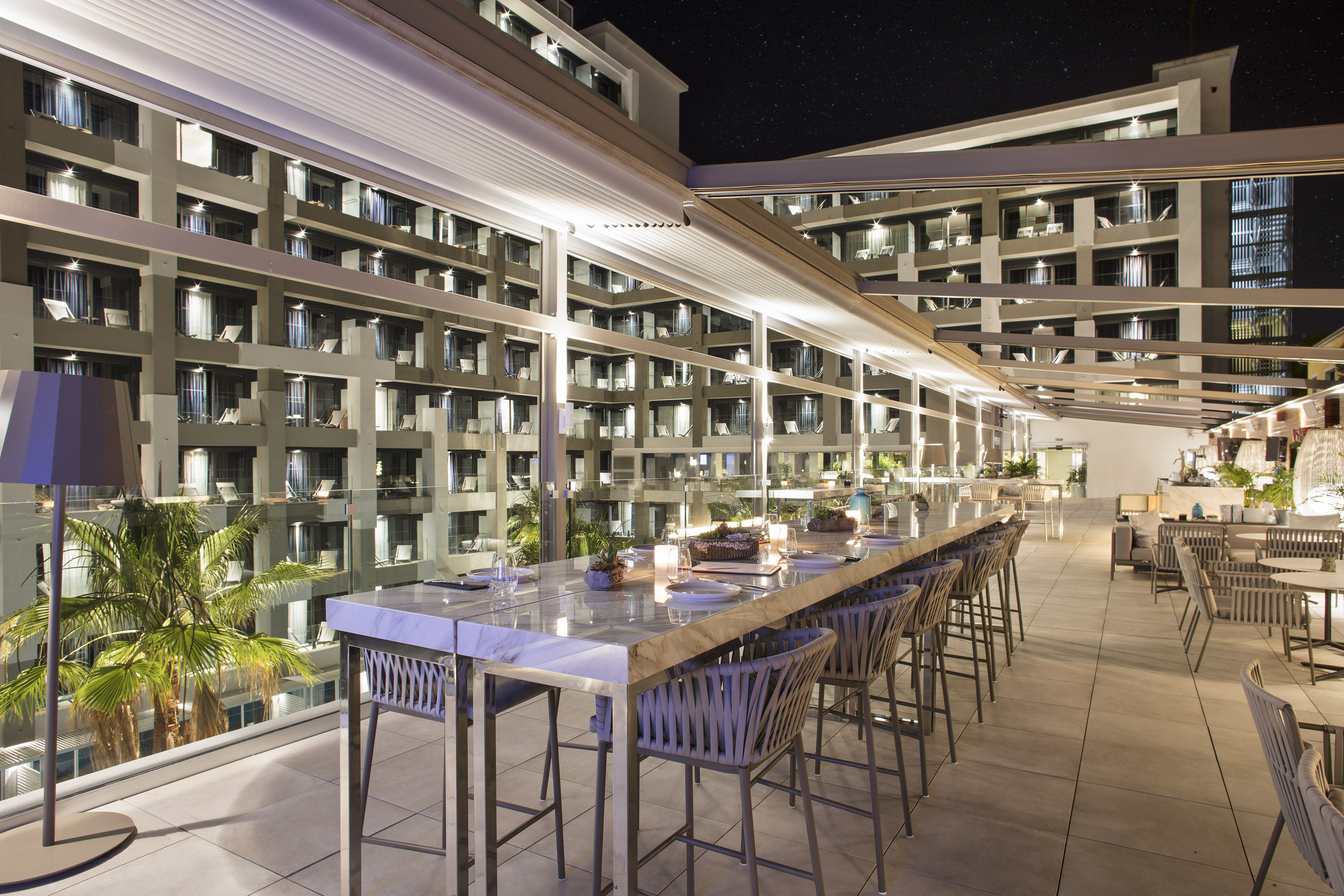 Modern rooftop bar with marble counter, bar stools, lit hotel at night.