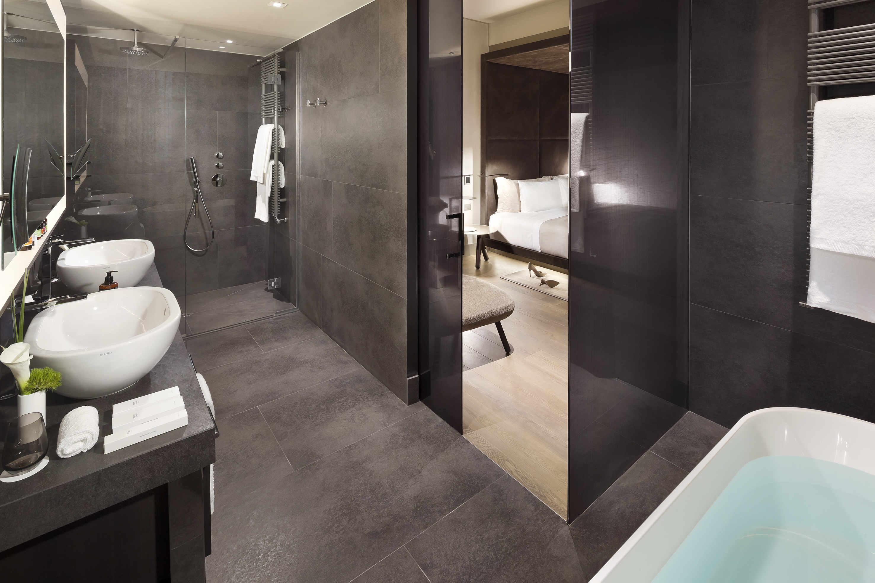 Dark tiled modern bathroom with double vanity, glass shower, and adjacent bedroom.