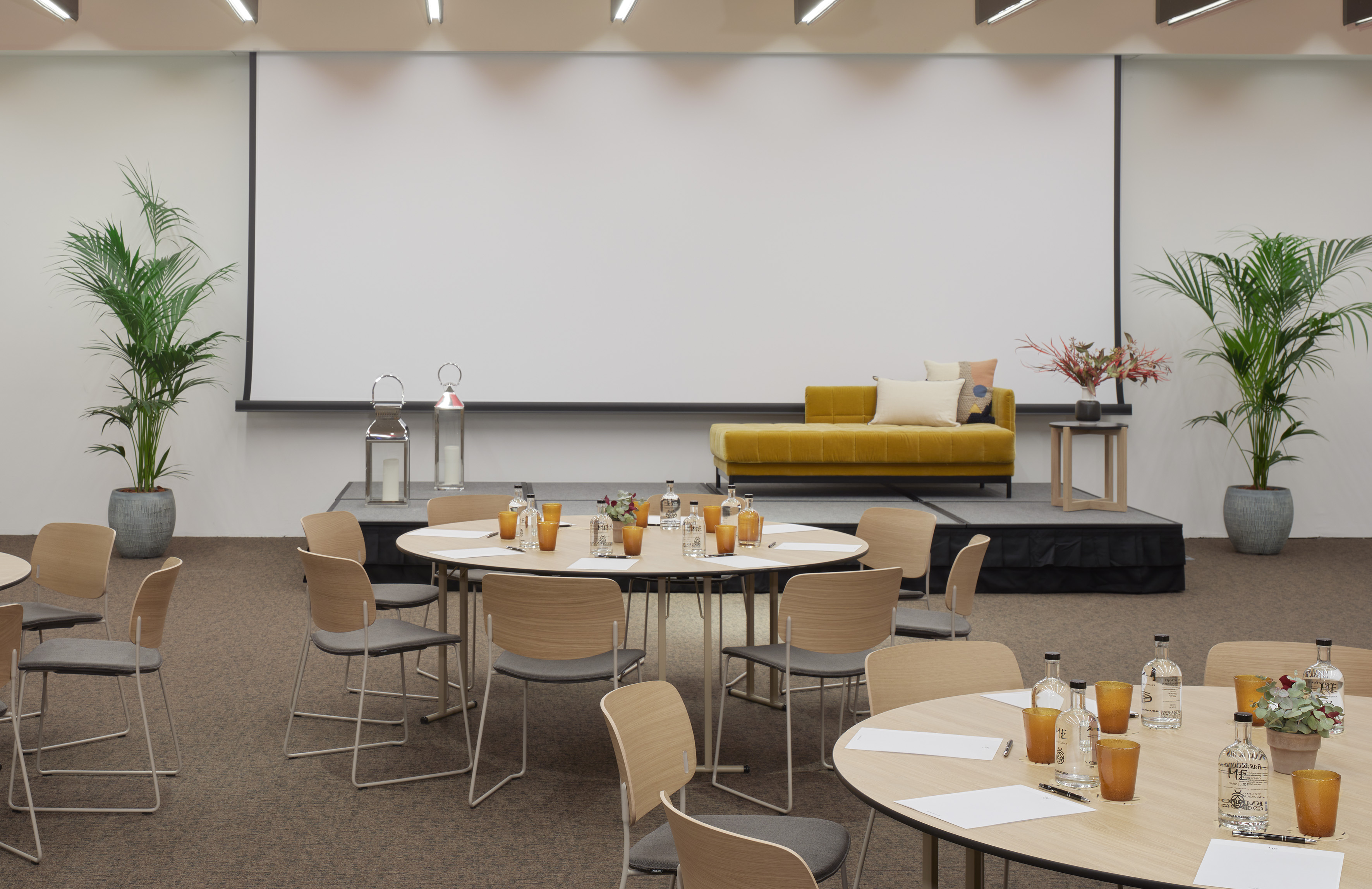 Conference room with round tables, screen, plants, and stage with yellow sofa.