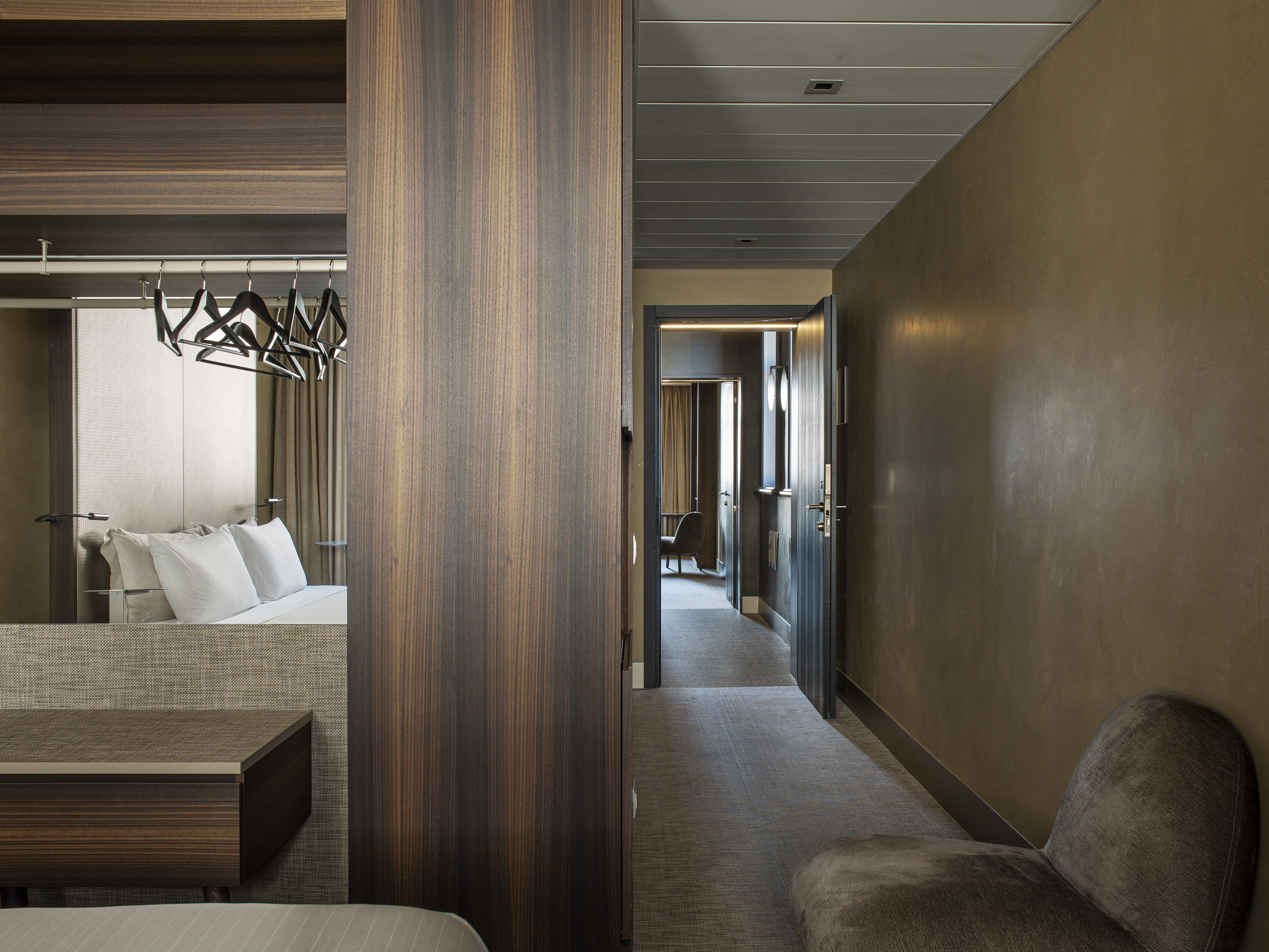 Minimalist hotel room with dark wood wardrobe, bed, and entrance hallway.