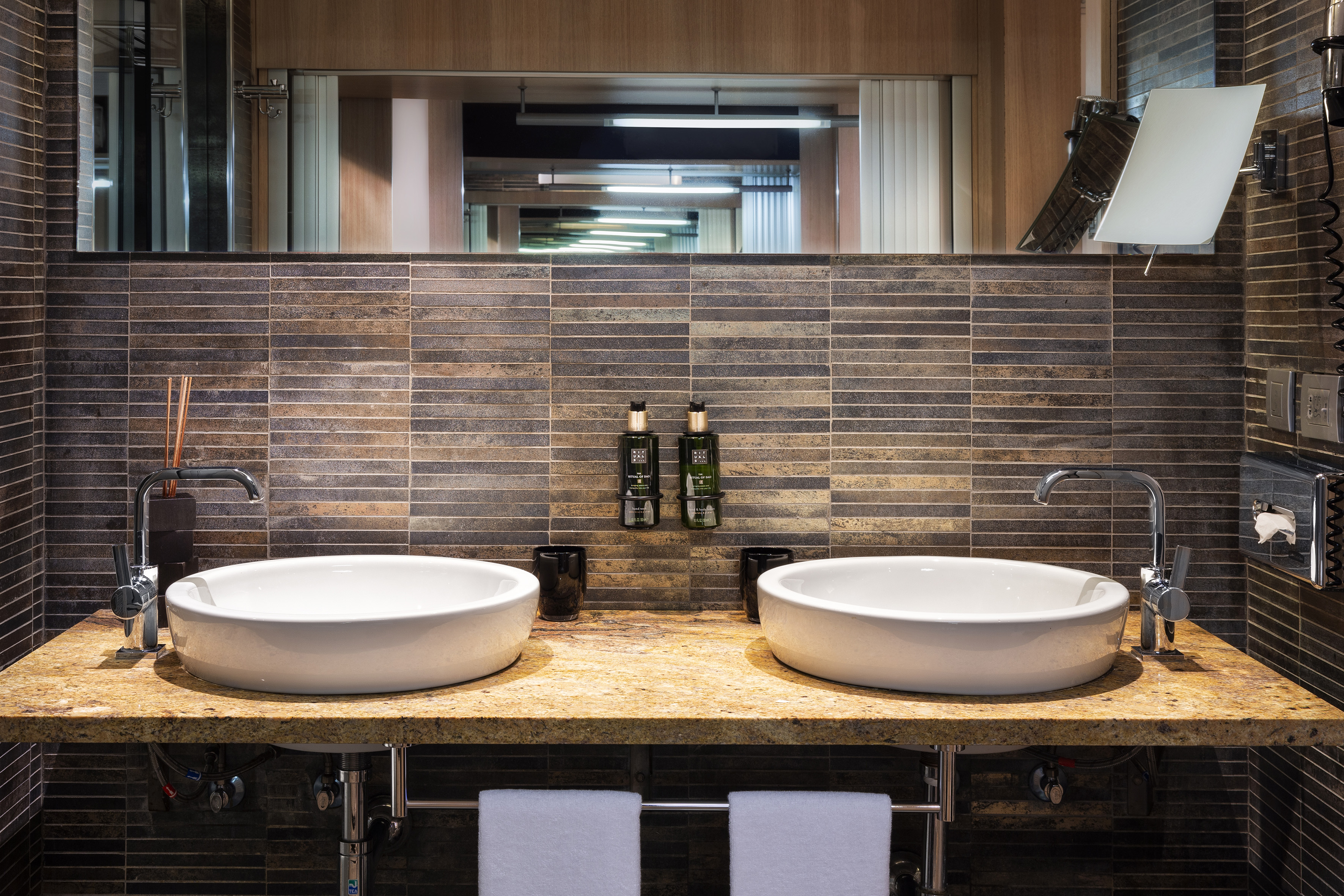Modern hotel bathroom with double vessel sinks, granite counter, and tiled wall.
