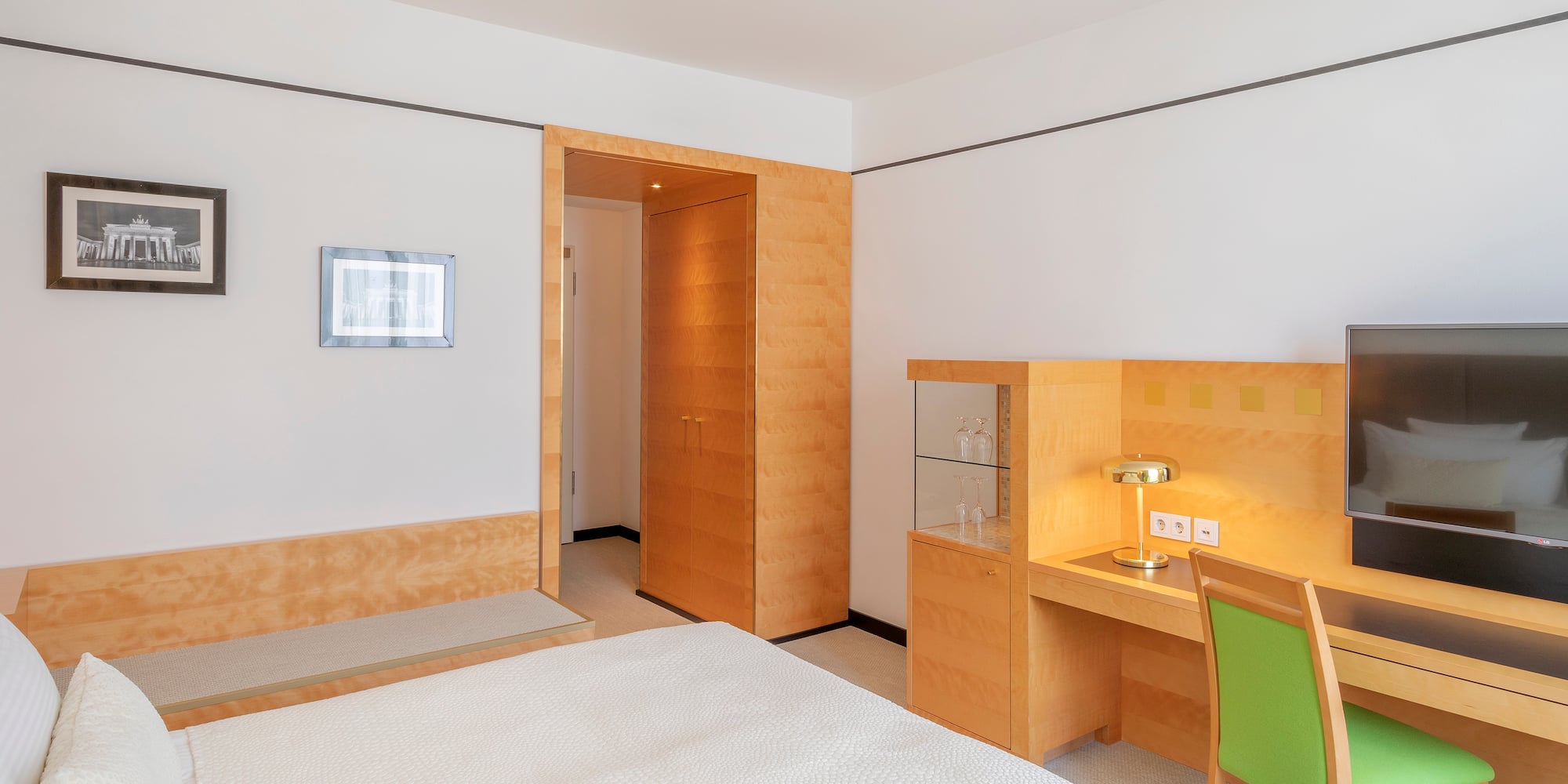 Clean hotel room with white bed, wooden desk, green chair, and TV.