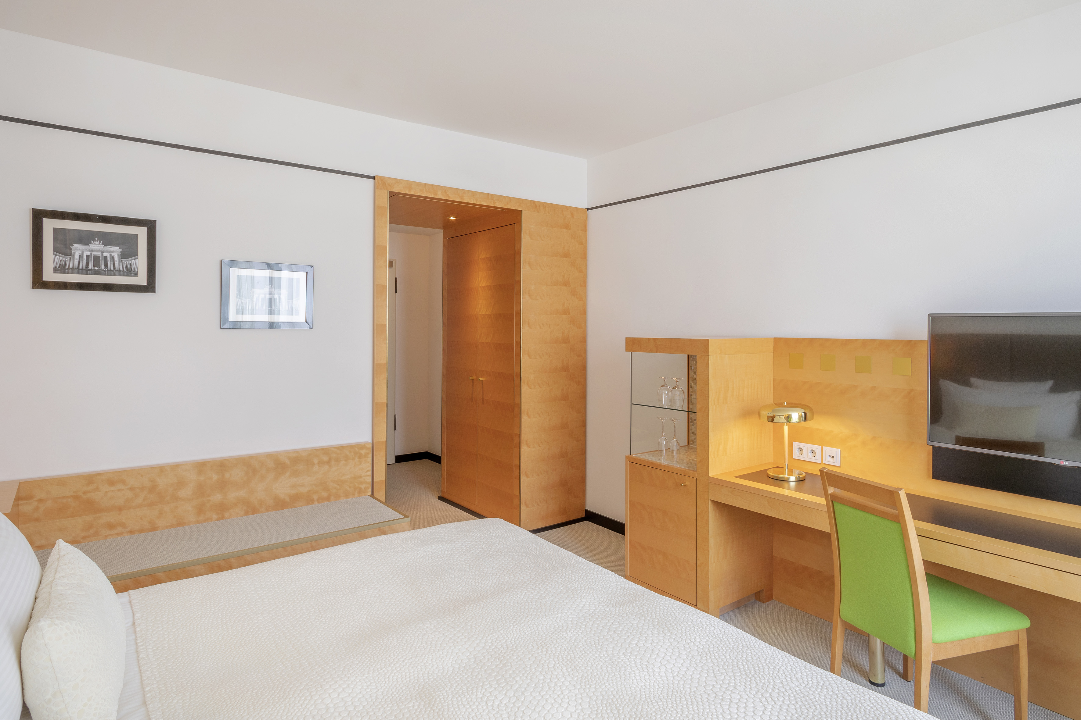 Clean hotel room with white bed, wooden desk, green chair, and TV.