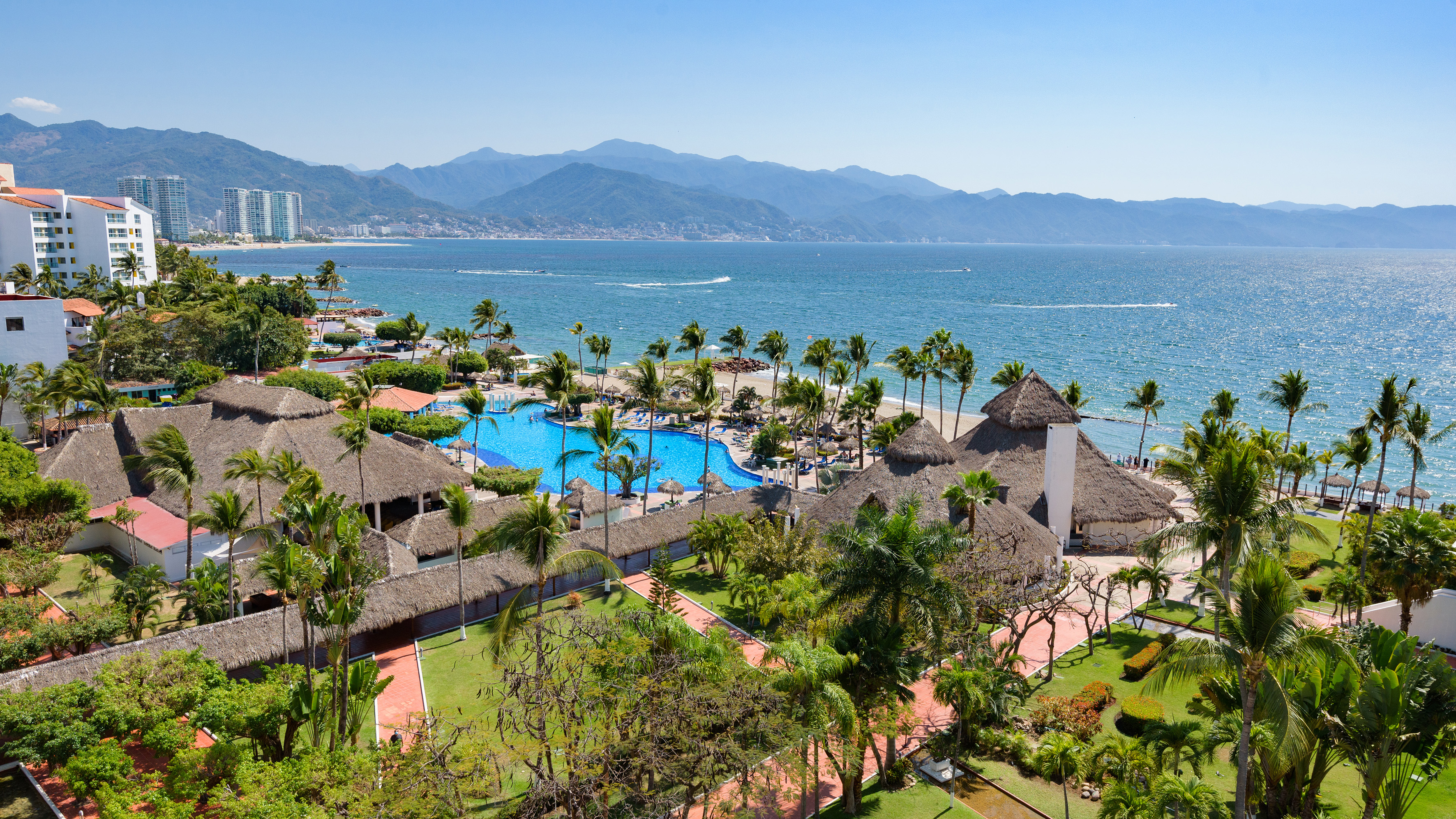 Beachfront tropical resort with large pool, palm trees, ocean, mountains.