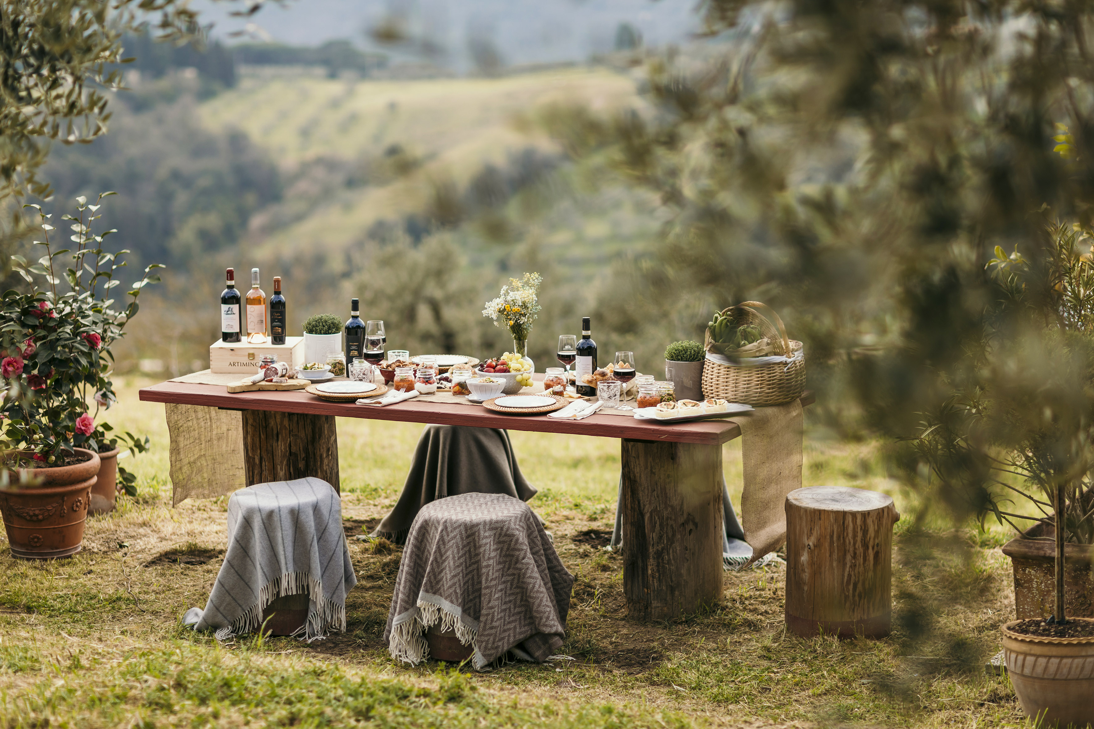 Al fresco dining table with wine, food, blankets, and scenic Tuscan landscape.
