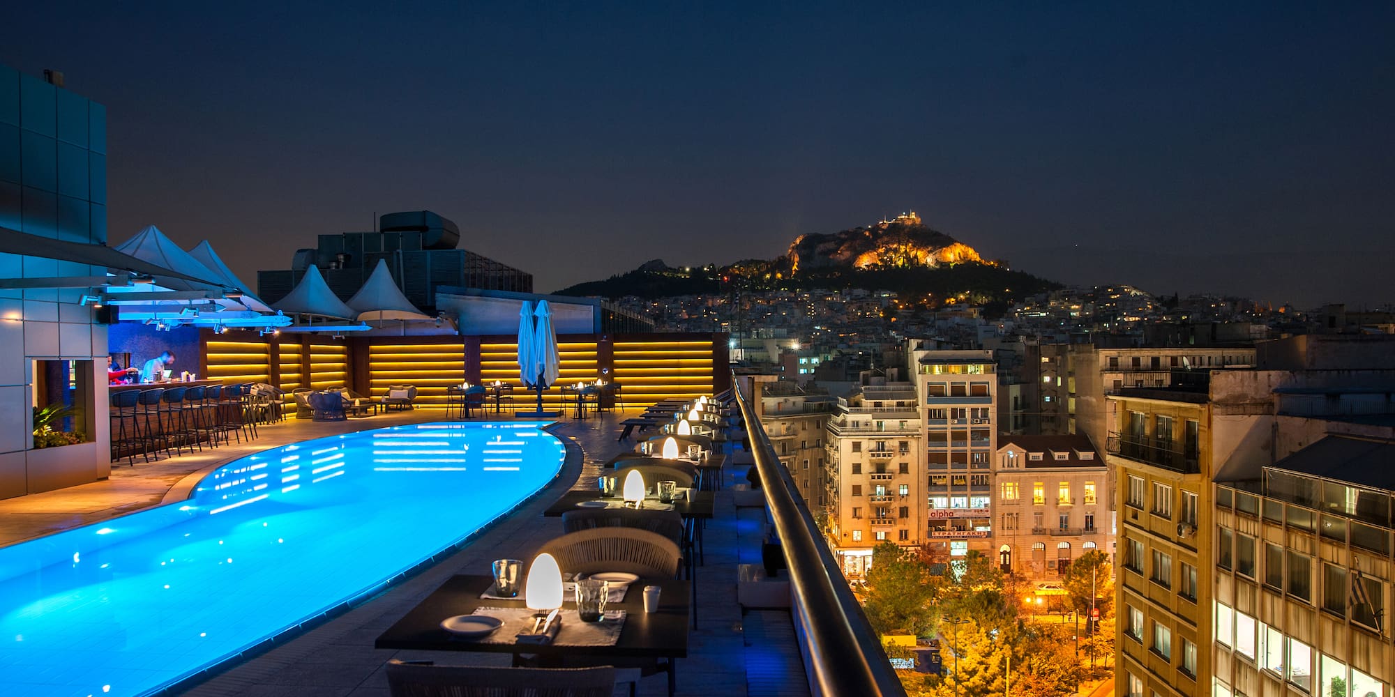 a rooftop pool with tables and chairs and a city in the background