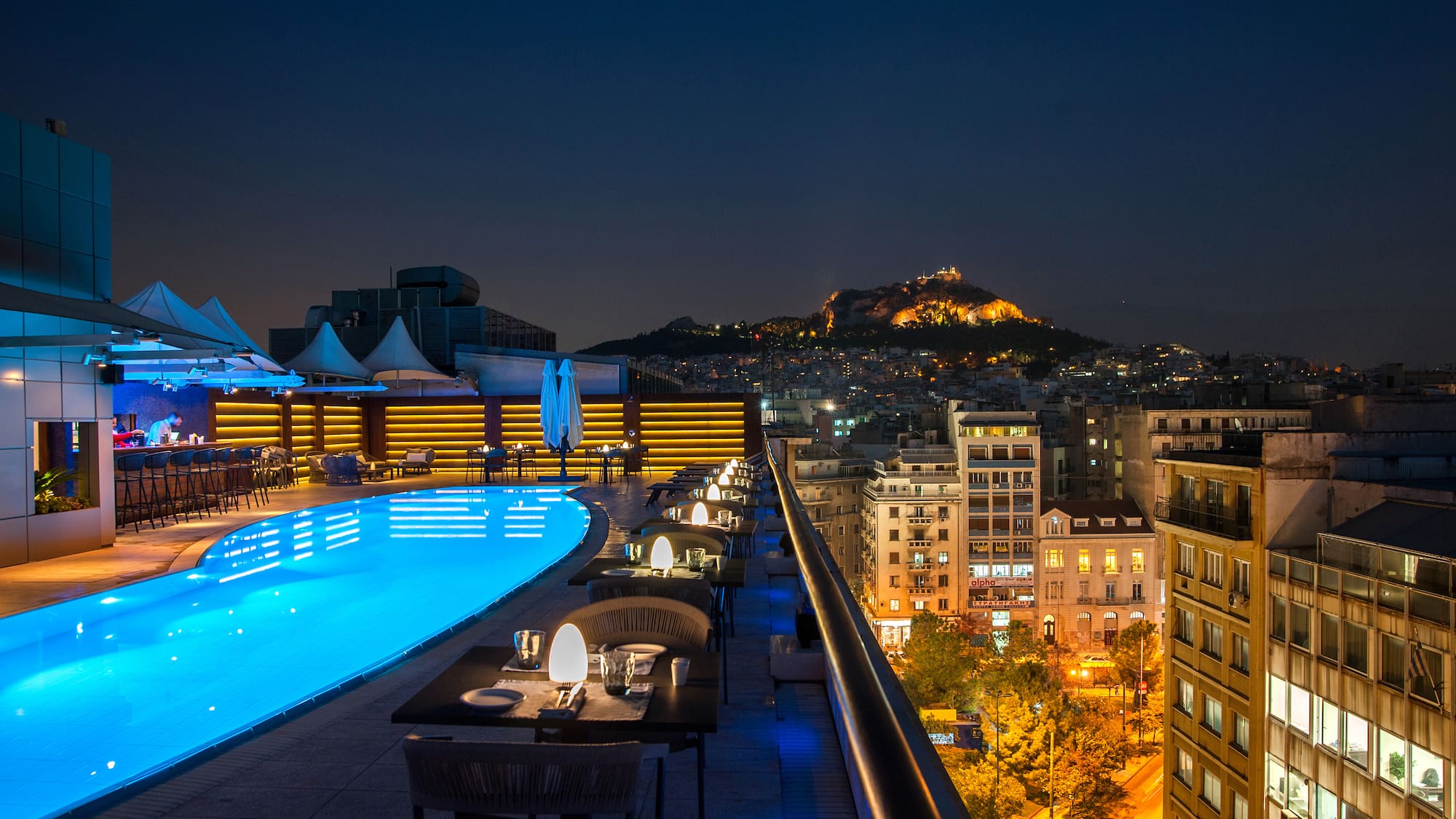 a rooftop pool with tables and chairs and a city in the background