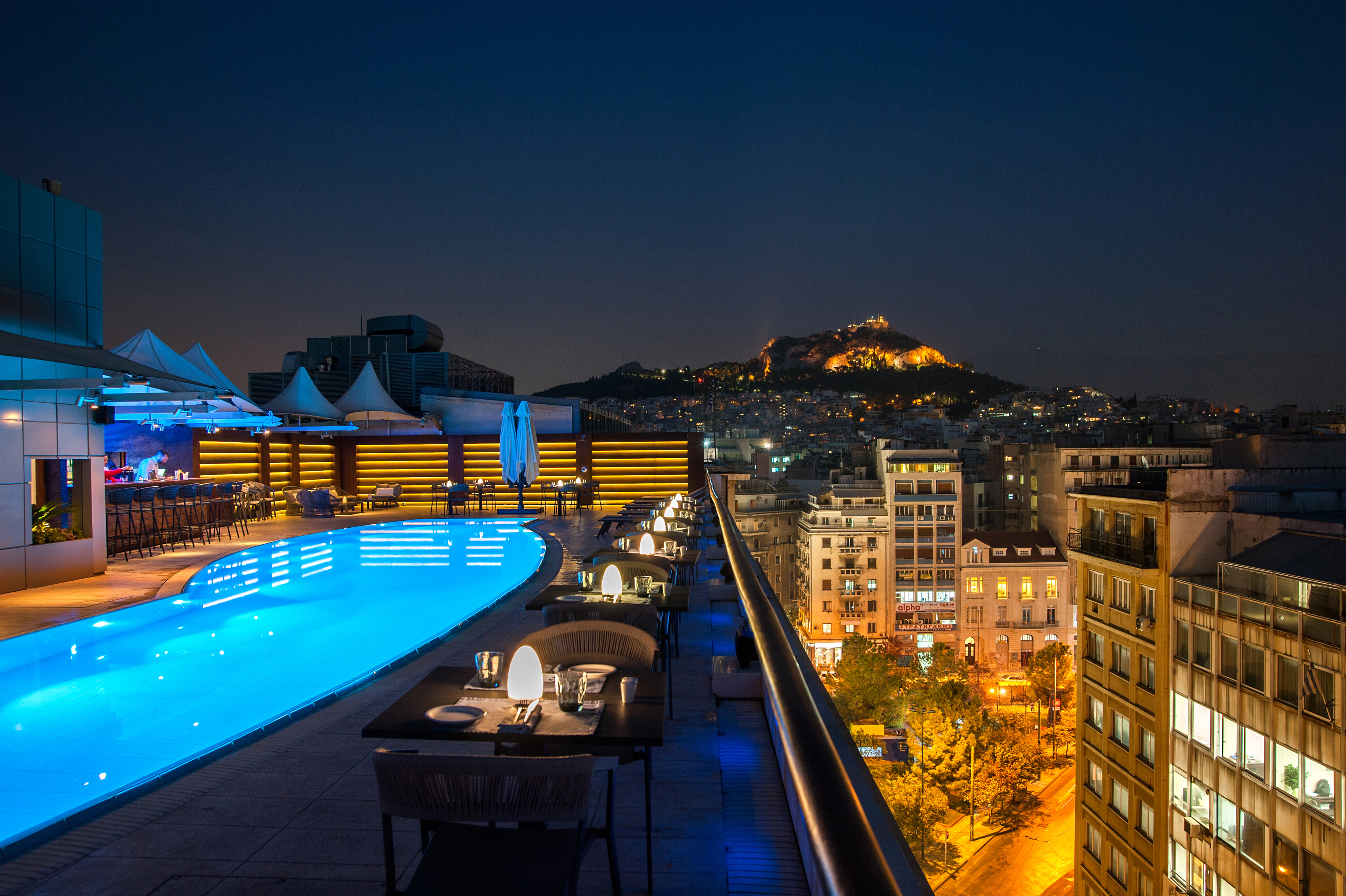 a rooftop pool with tables and chairs and a city in the background