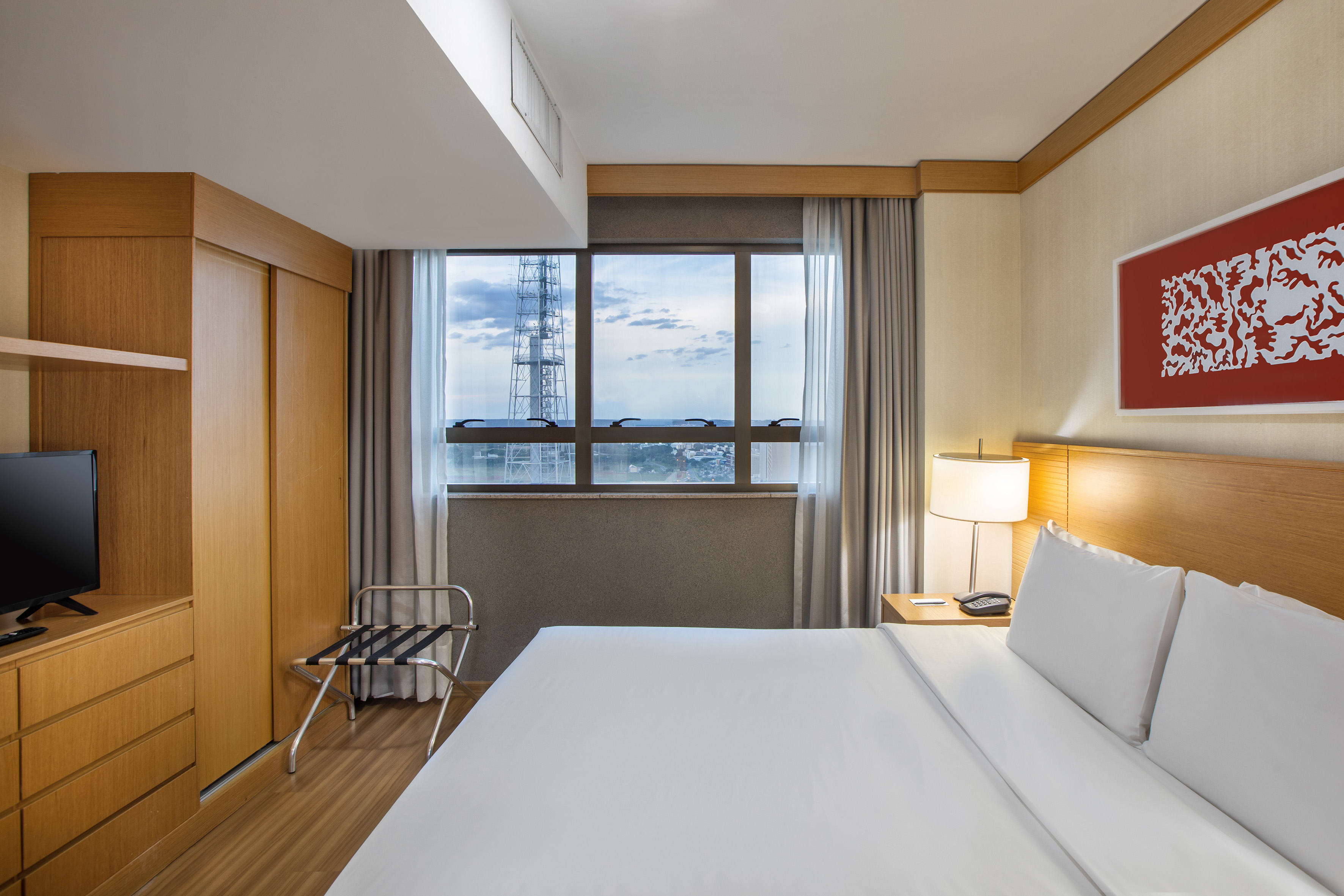 Modern hotel room with king bed, city view window, and wooden furniture.