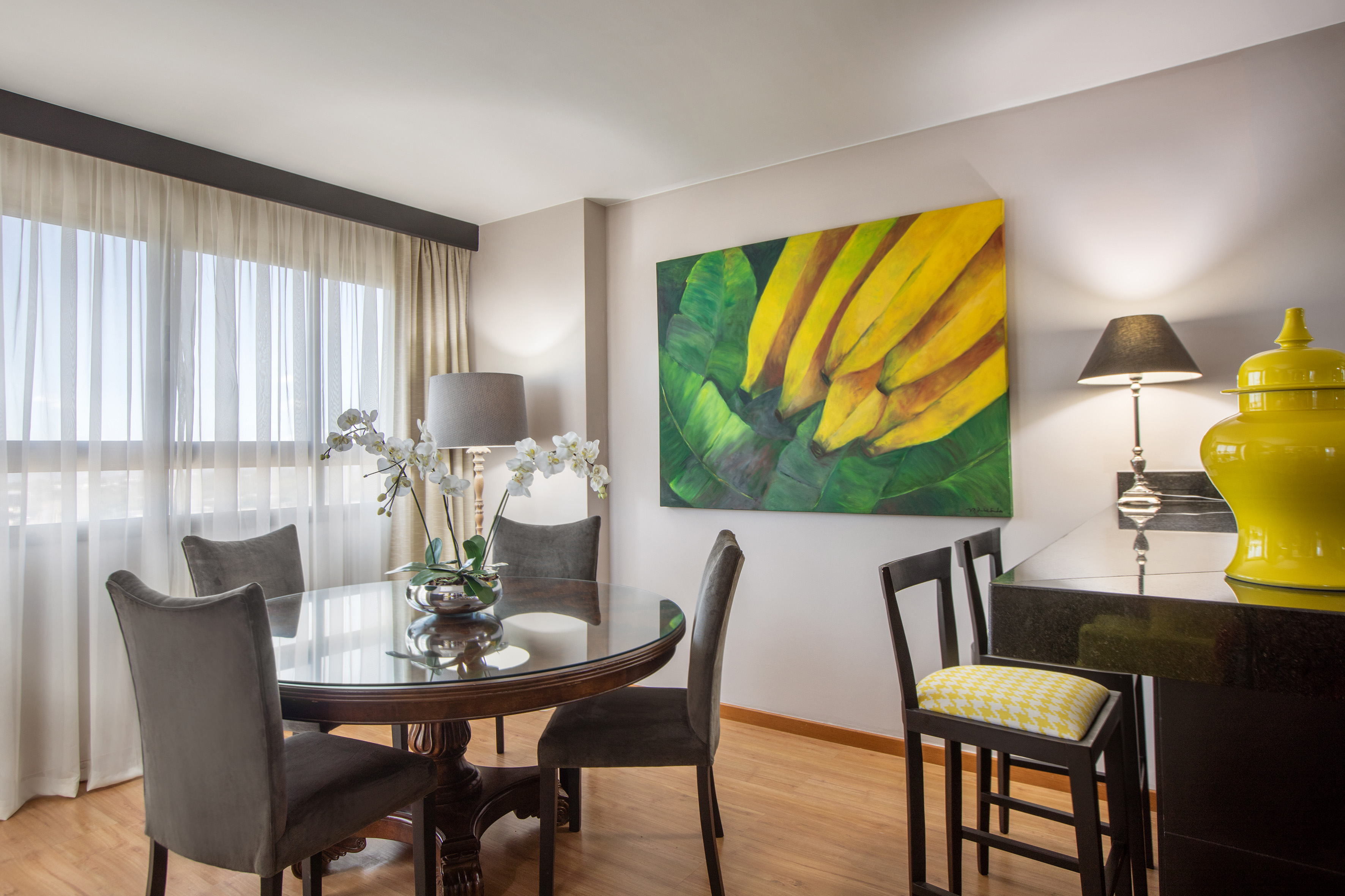 Dining area with round glass table, gray chairs, vibrant banana artwork.