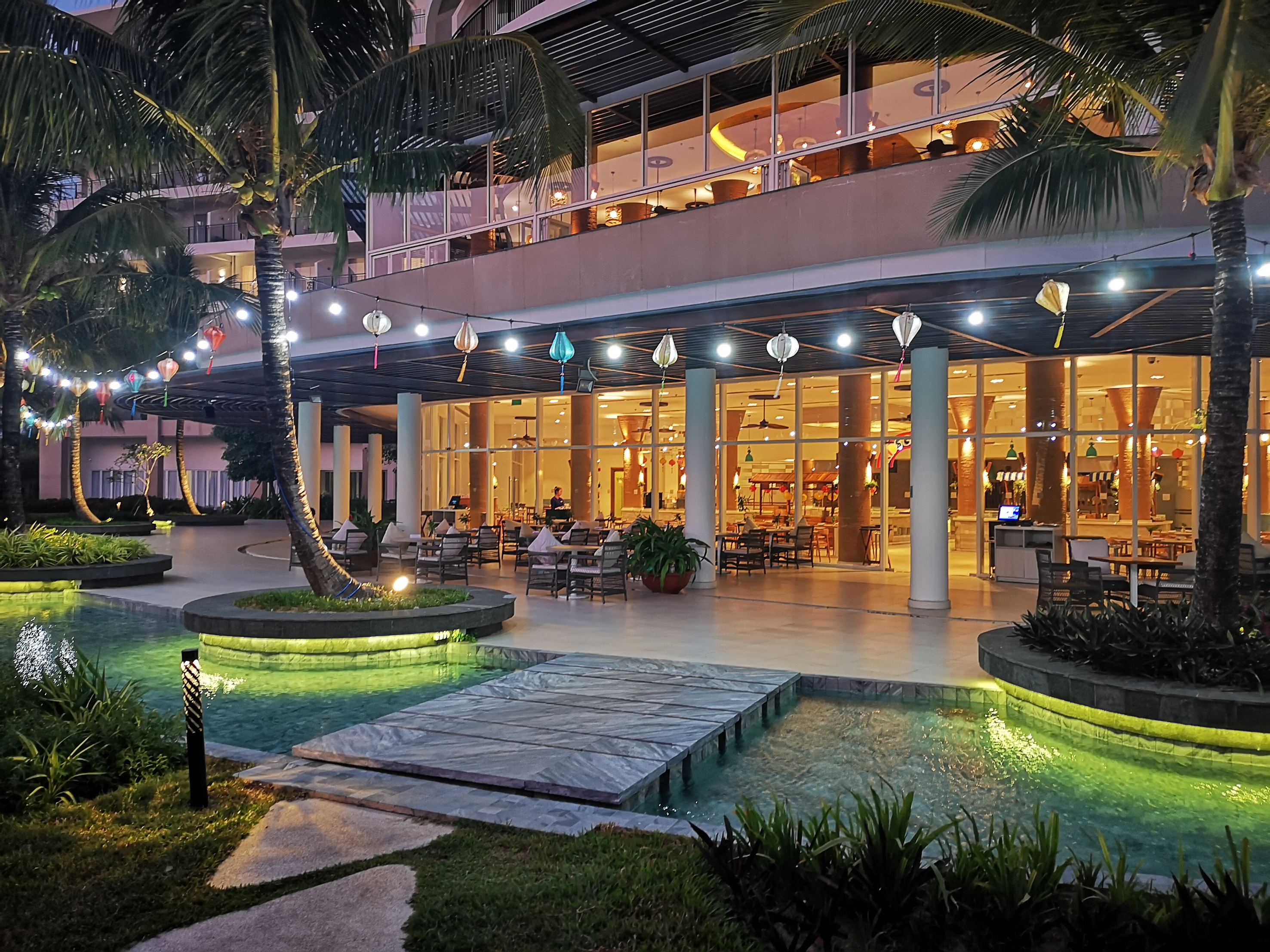 Evening resort patio, palm trees, festive lanterns, illuminated water features.