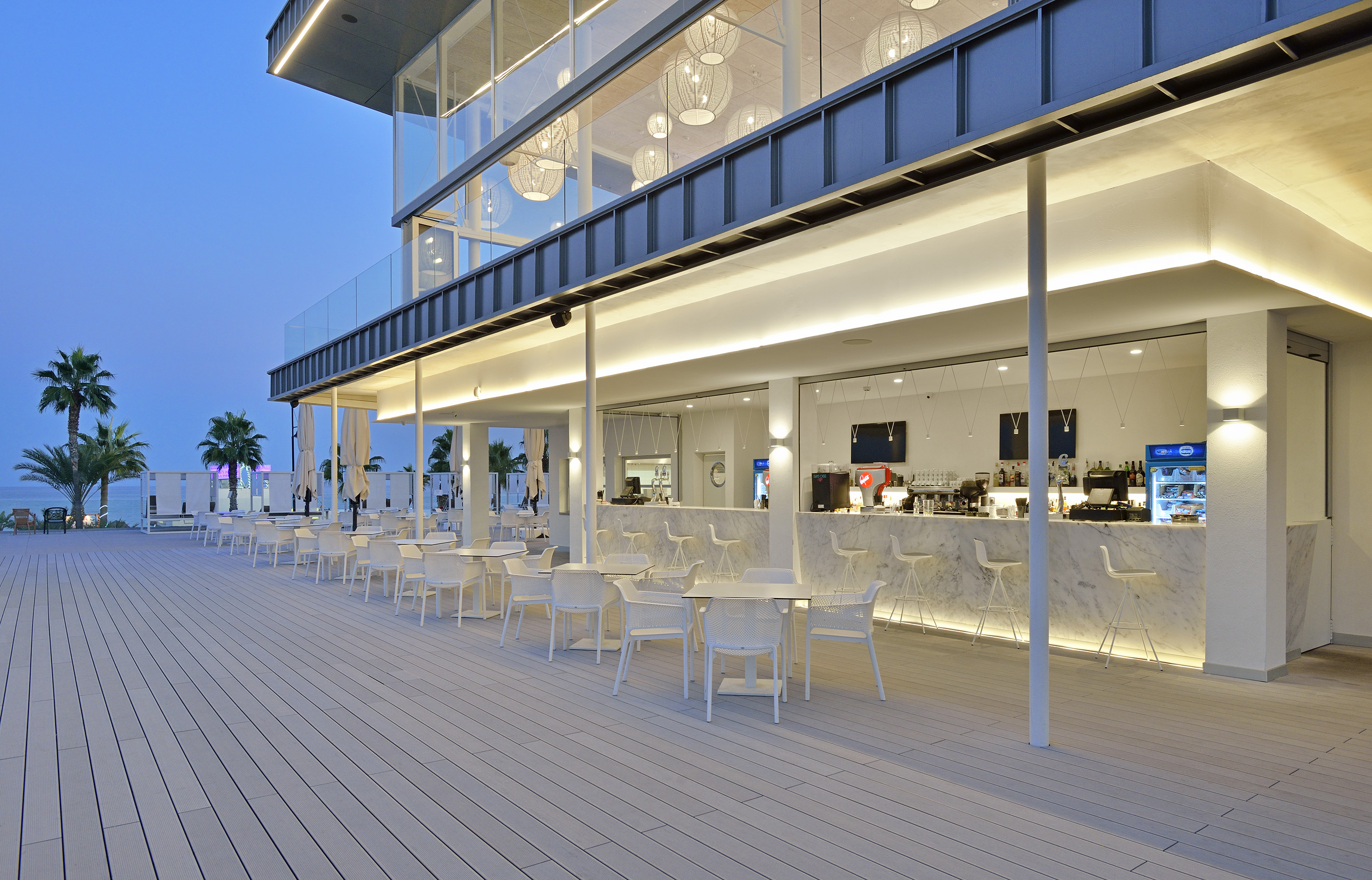 Modern beach club outdoor bar, white furniture, illuminated terrace, sea view.