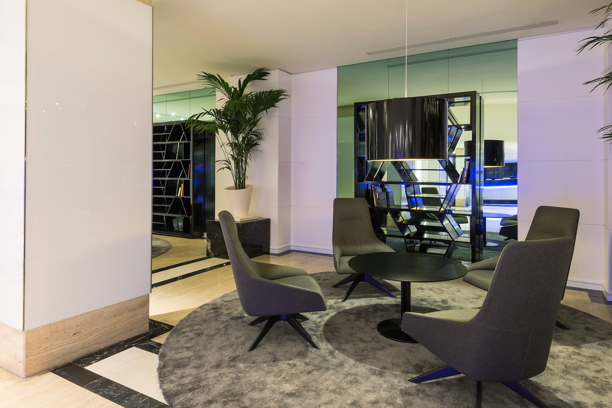 Modern hotel lobby lounge area with grey seating, round table, and palm tree.