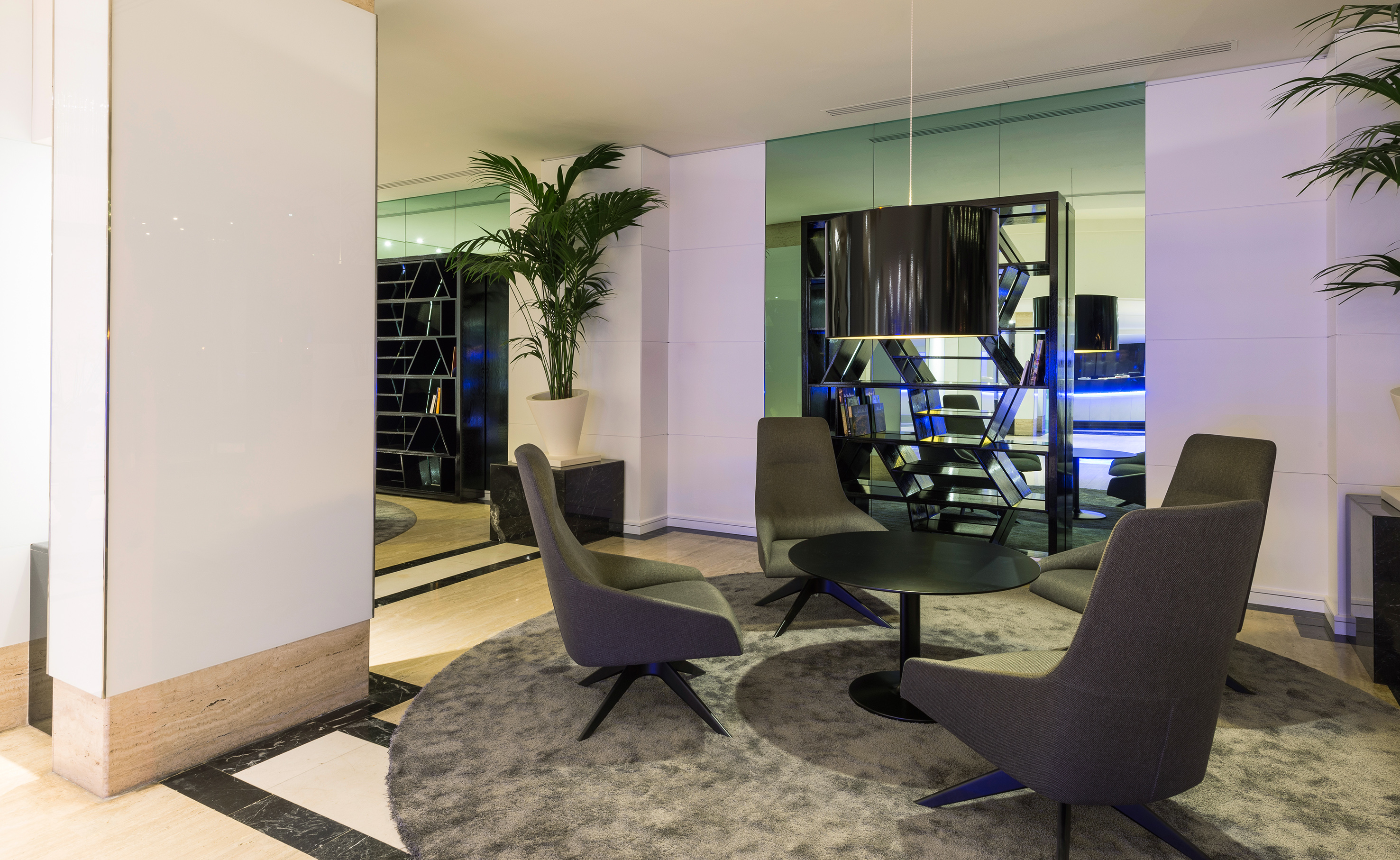 Modern hotel lobby lounge area with grey seating, round table, and palm tree.