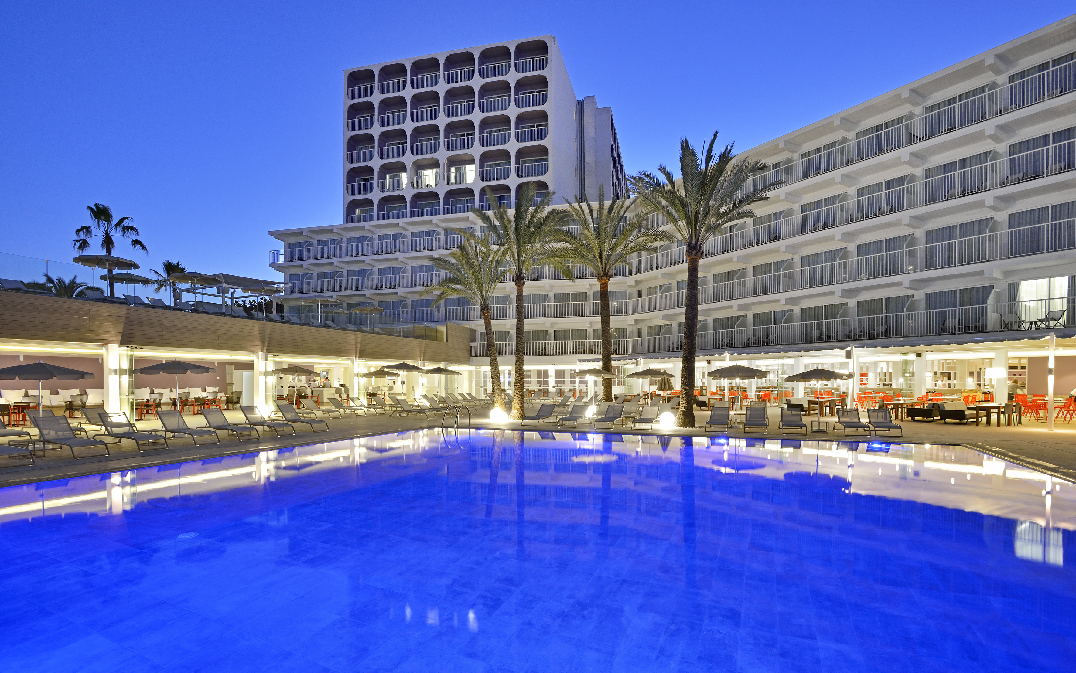 Modern hotel resort, illuminated blue pool, palm trees, loungers at night.