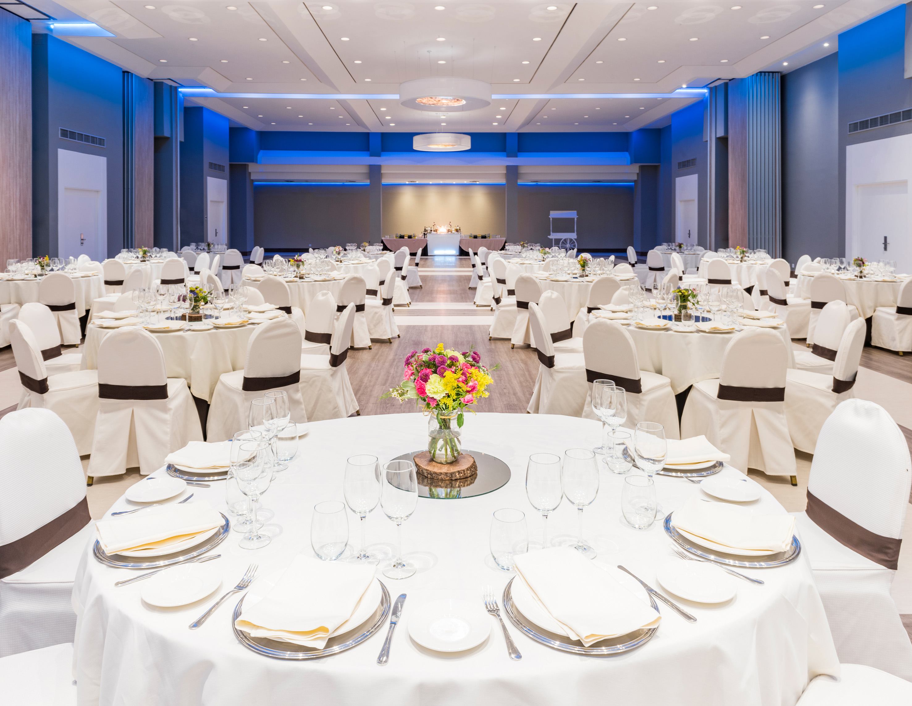 Elegant ballroom with white set tables, chairs, floral decor, and blue lighting.