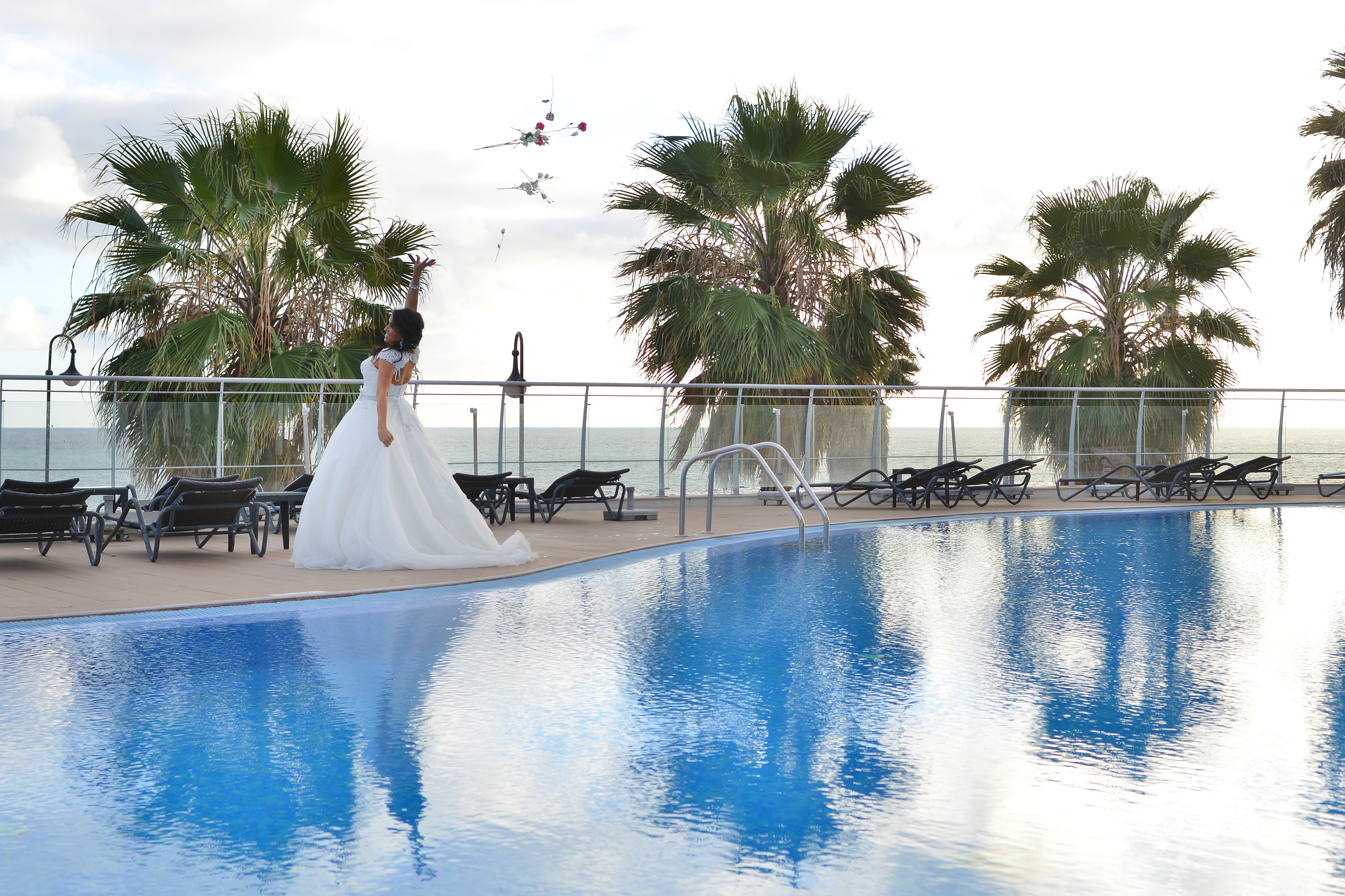 Bride in wedding dress tossing petals by resort pool with palm trees, ocean.