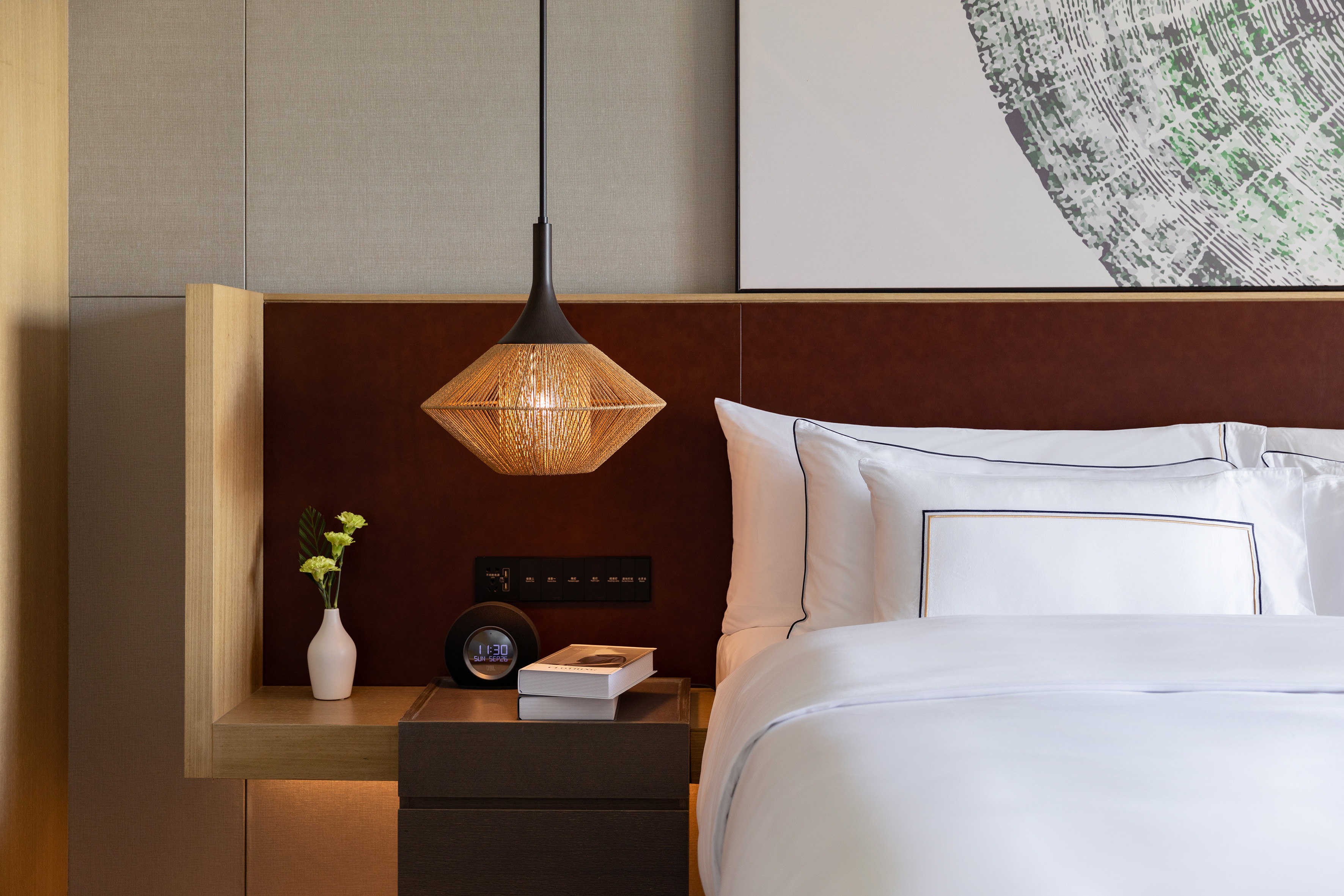 Modern hotel bedroom with white bedding, woven pendant light, and dark headboard.