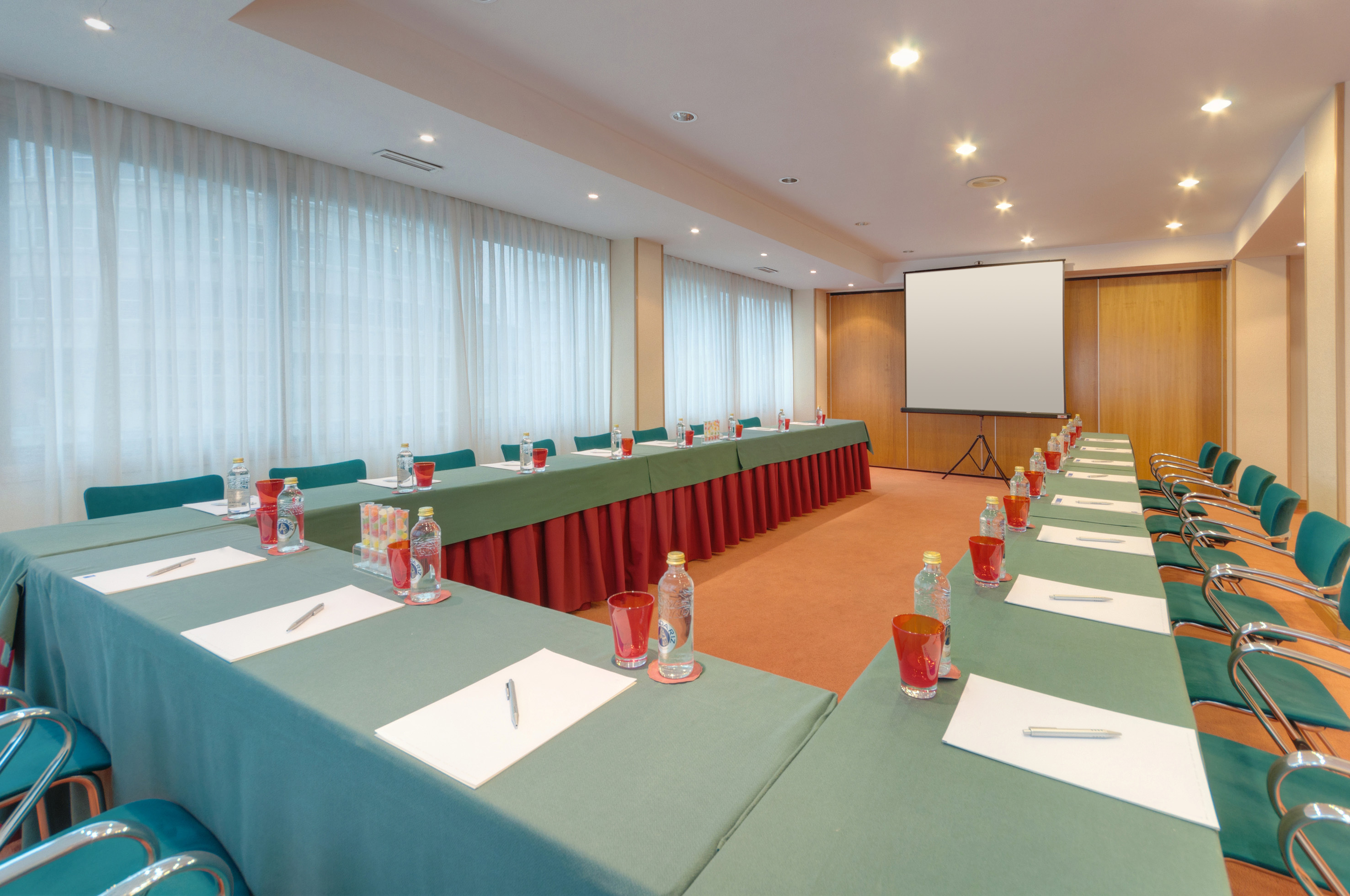 U-shaped conference room with green tables, chairs, water, and a projector screen.