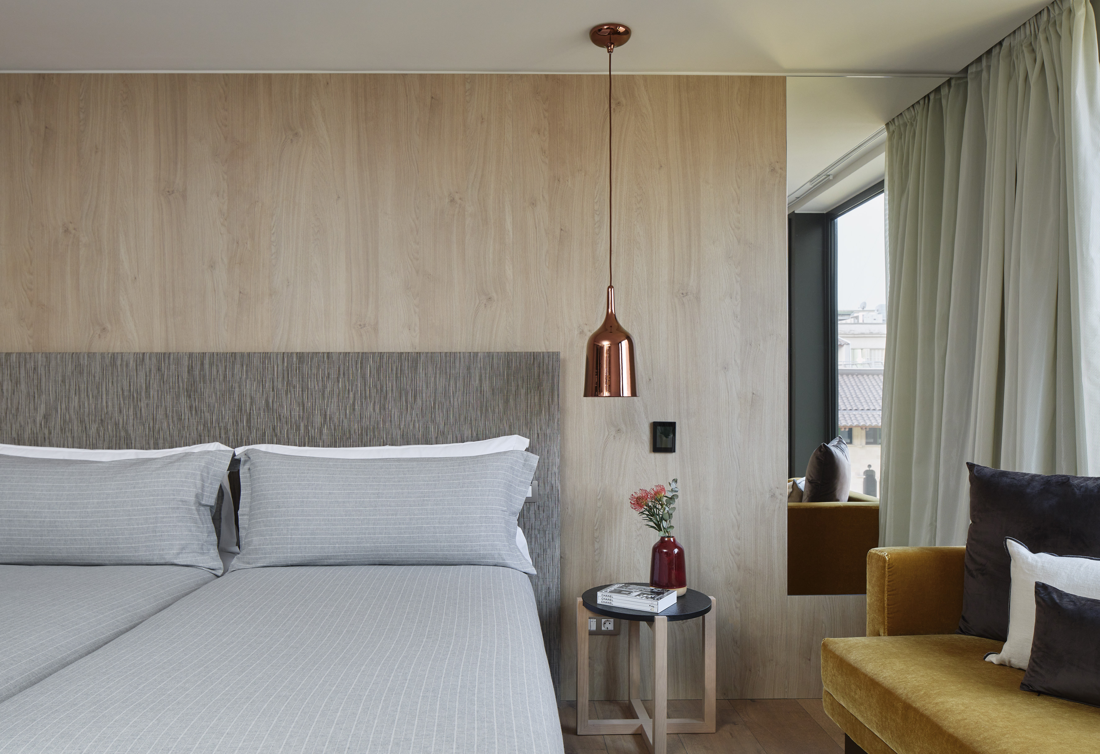 Modern hotel bedroom, grey bed, wood paneling, copper pendant light, velvet sofa.