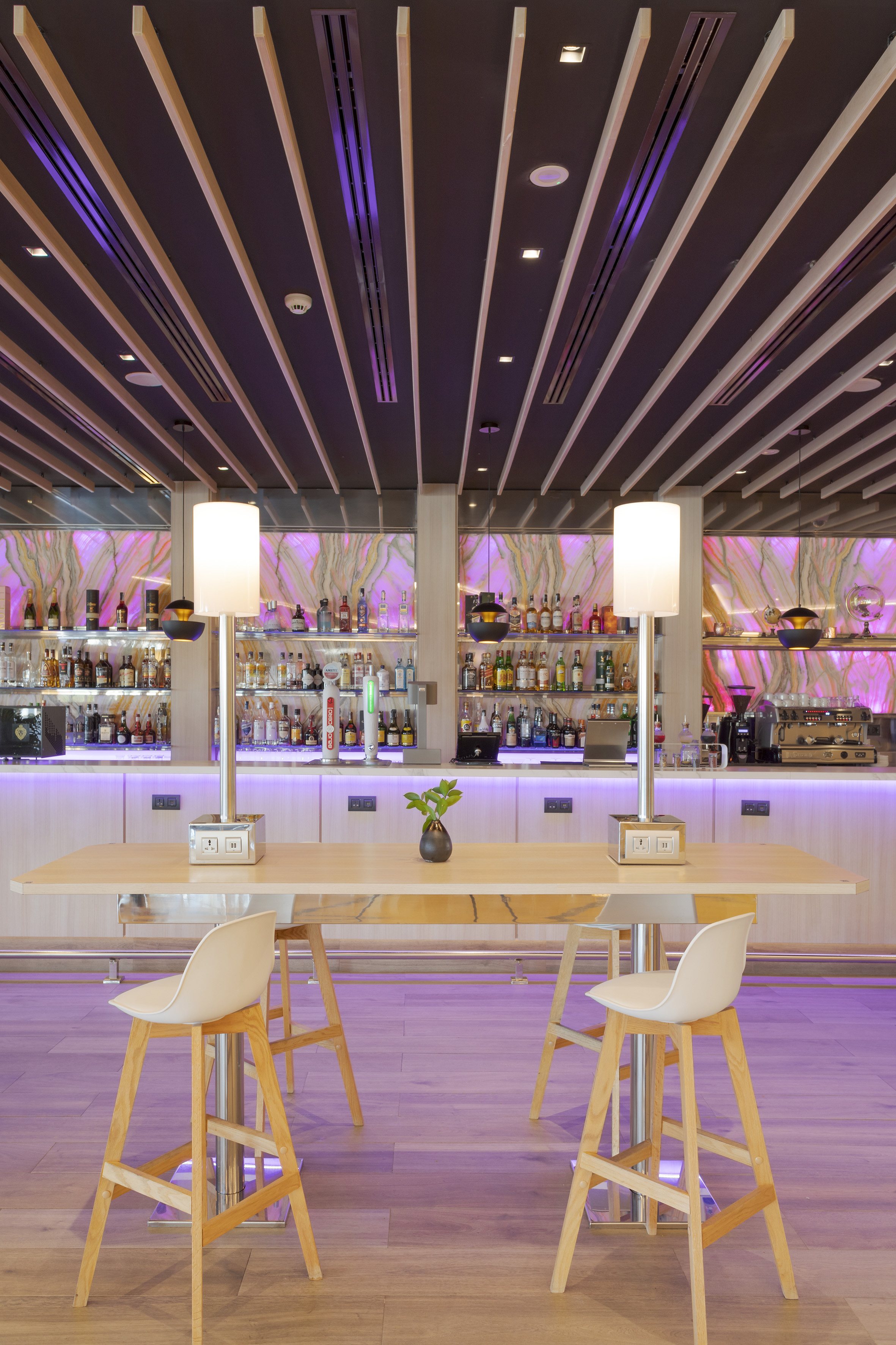 Modern bar with purple lighting, high table, white stools, and liquor shelves.