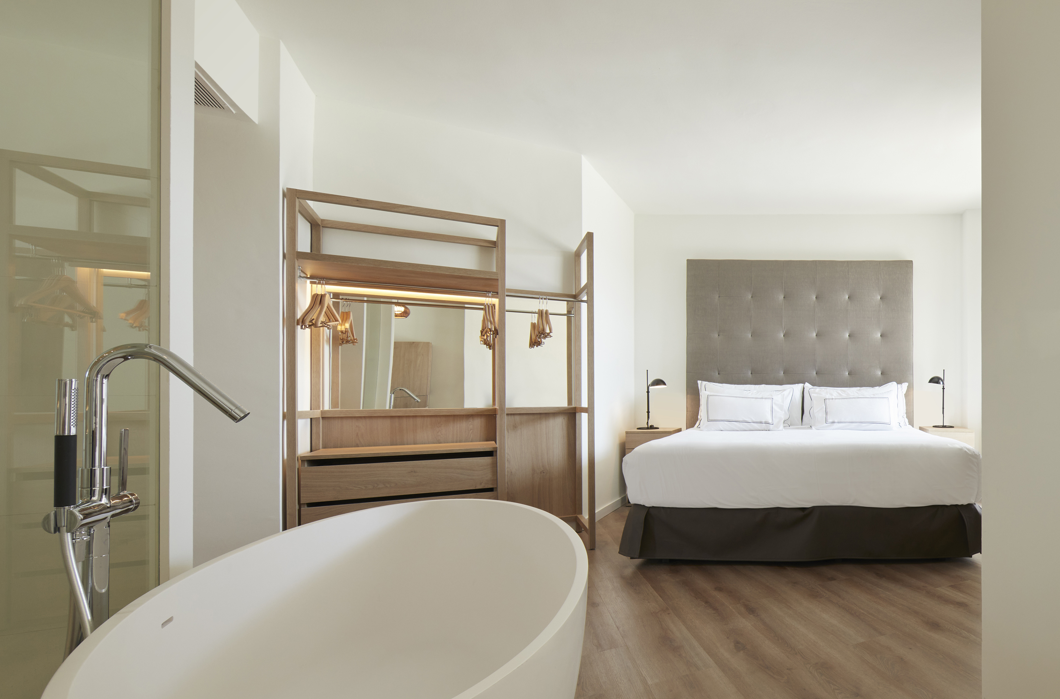 Modern hotel bedroom with white freestanding bathtub, king bed, and open wood closet.