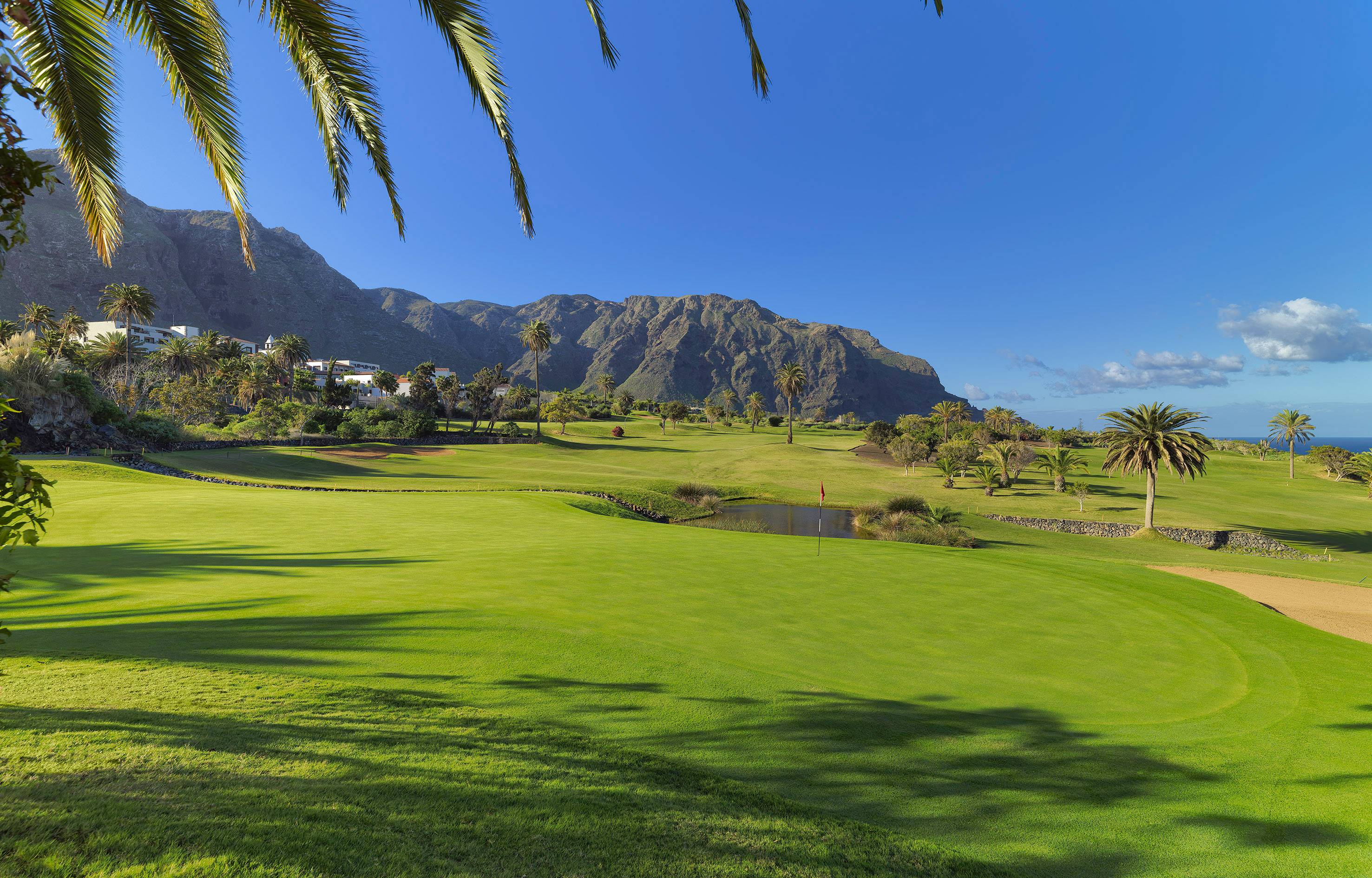 Green golf course with palm trees, mountains, and a pond under blue sky.