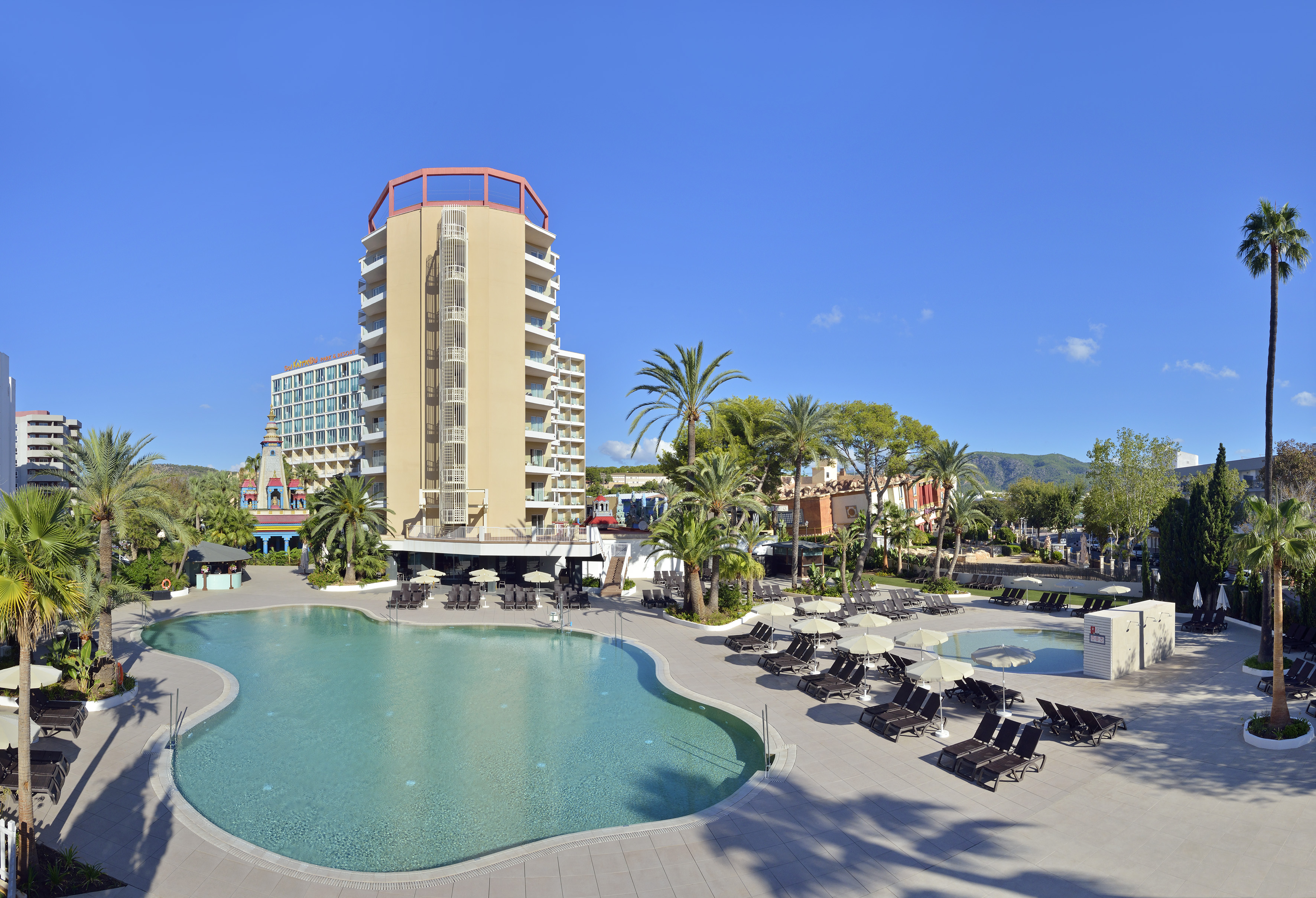 Sunny hotel resort with large outdoor pool, palm trees, and sun loungers.
