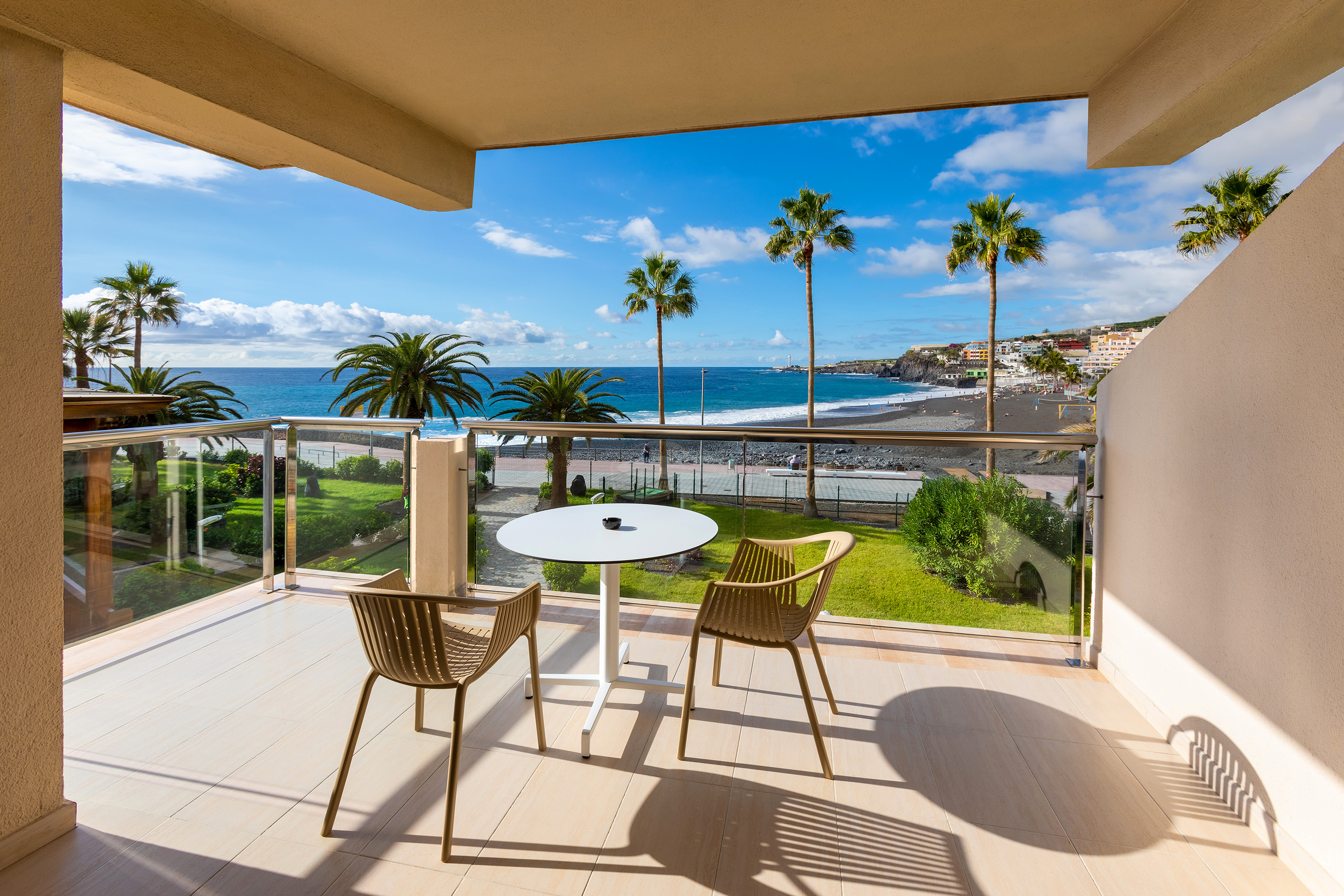 Sunny balcony with ocean view, palm trees, and a black sand beach.