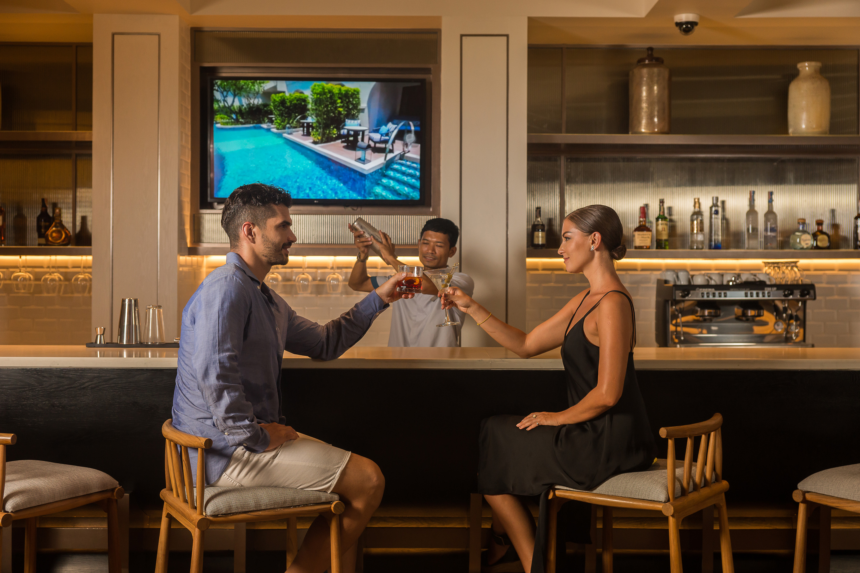 Couple toasting cocktails at a resort bar, bartender mixes drinks.