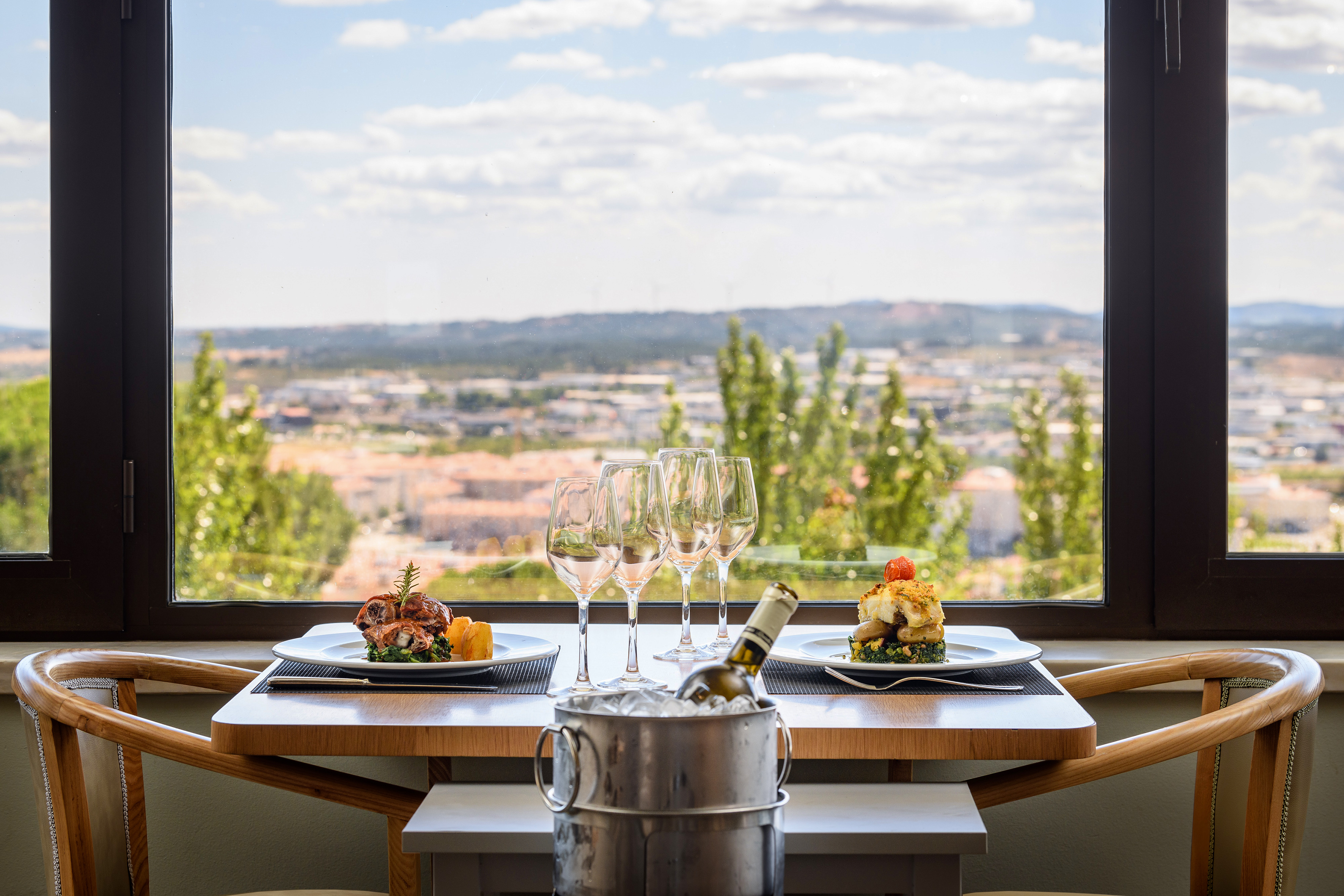 Gourmet restaurant dining table with two meals, wine, and panoramic city view.