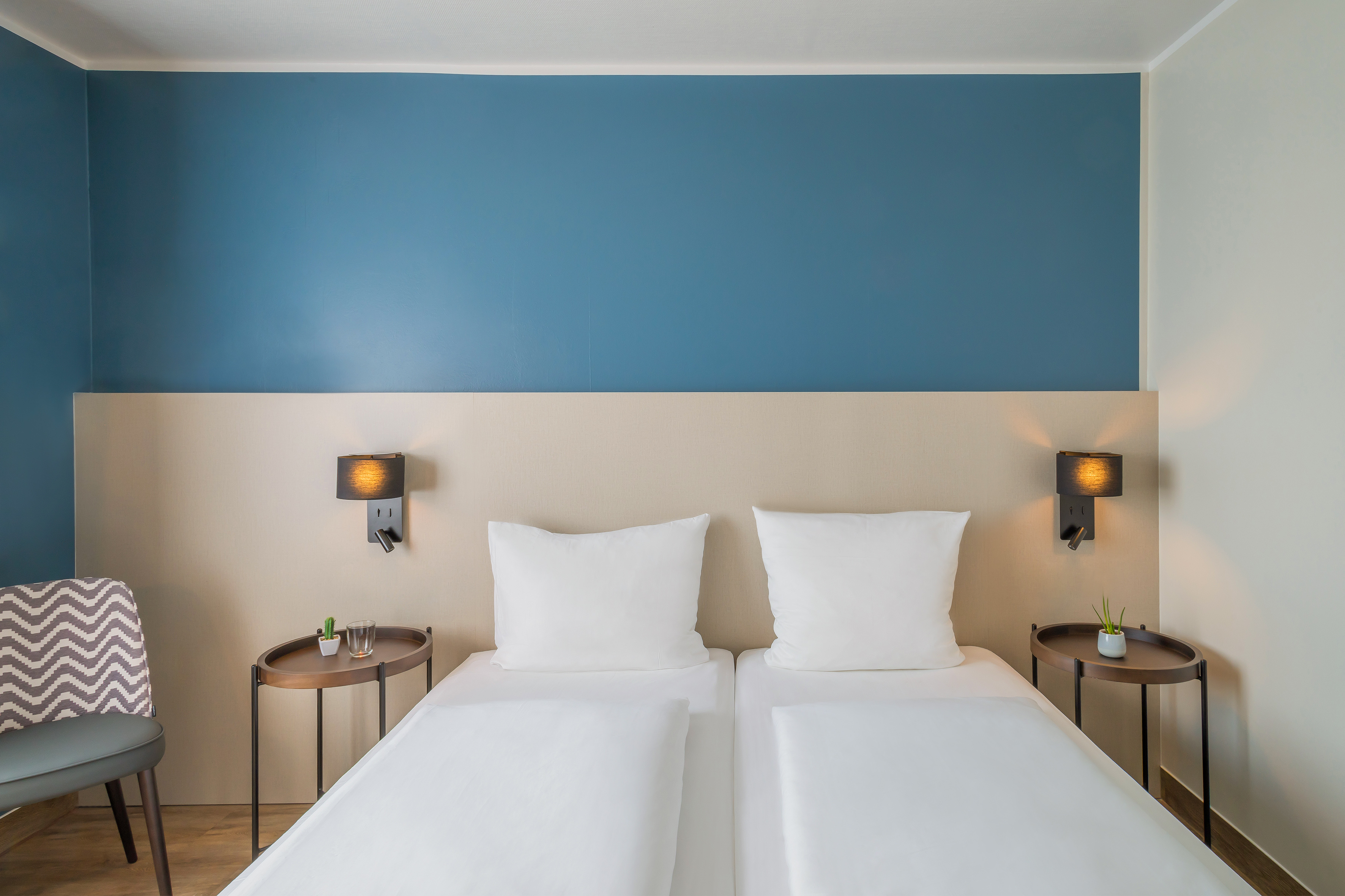 Modern hotel room with twin beds, blue accent wall, and warm bedside lamps.