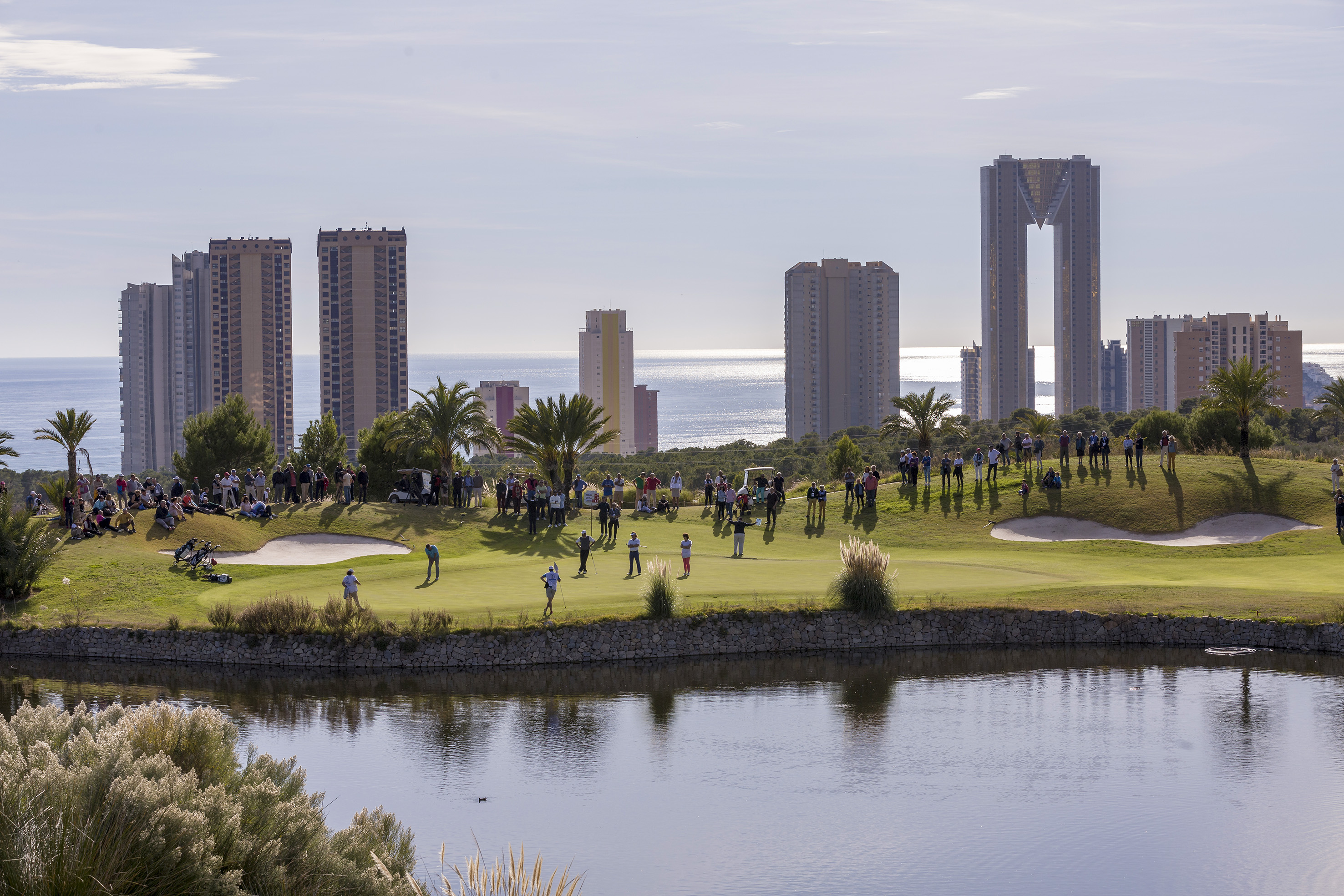 Benidorm golf course with players, spectators, water hazard, and coastal skyline.