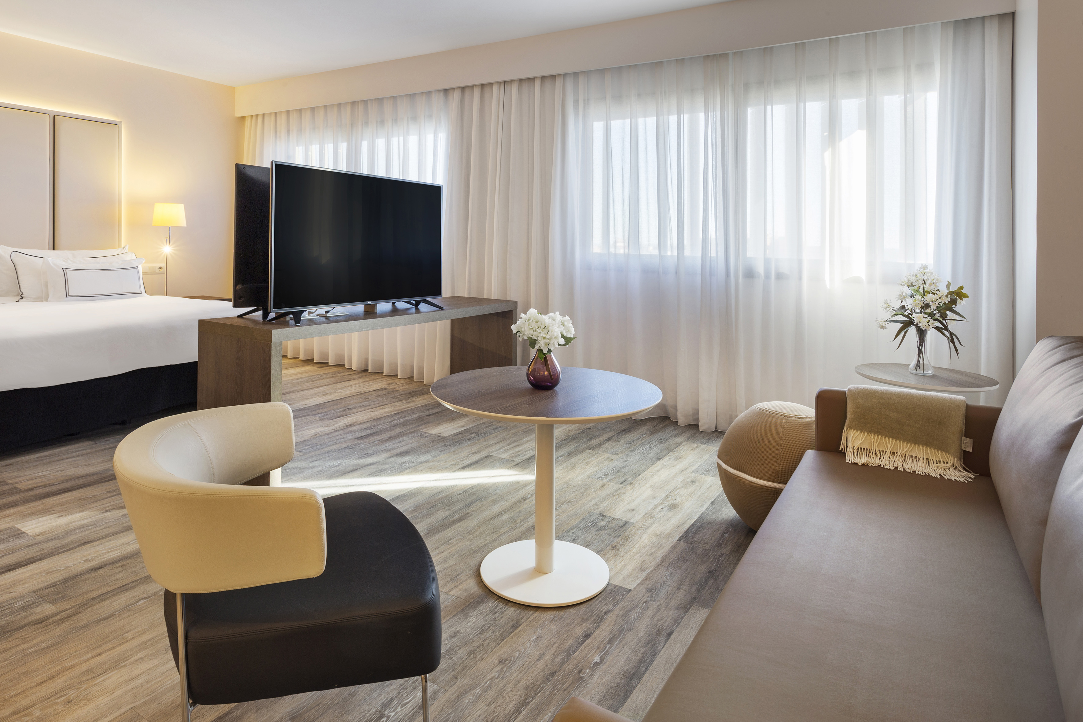 Modern hotel suite with bed, TV, sofa, and bright windows with sheer curtains.