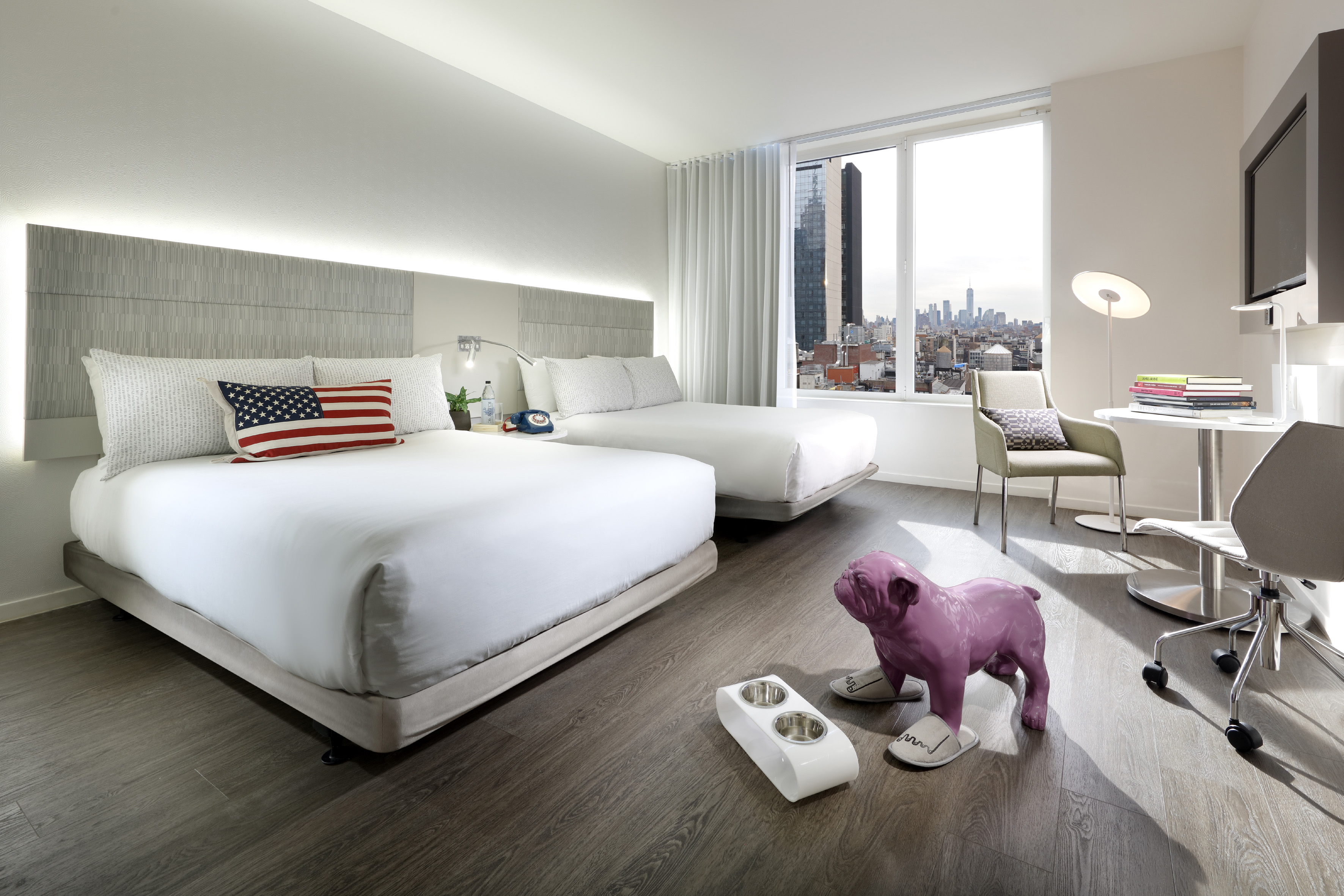 Pet-friendly hotel room with two beds, American flag pillow, and city view.
