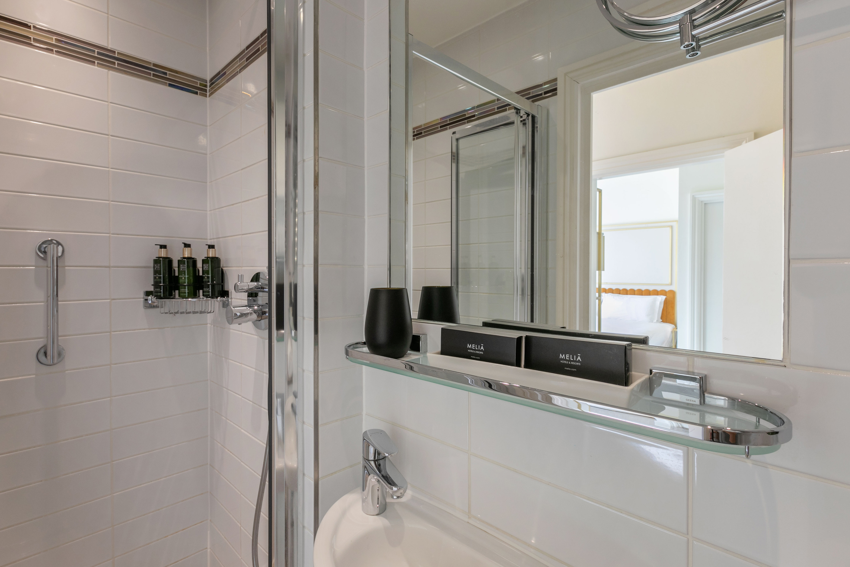 Clean hotel bathroom with white tiled shower, amenities, and bedroom reflection.