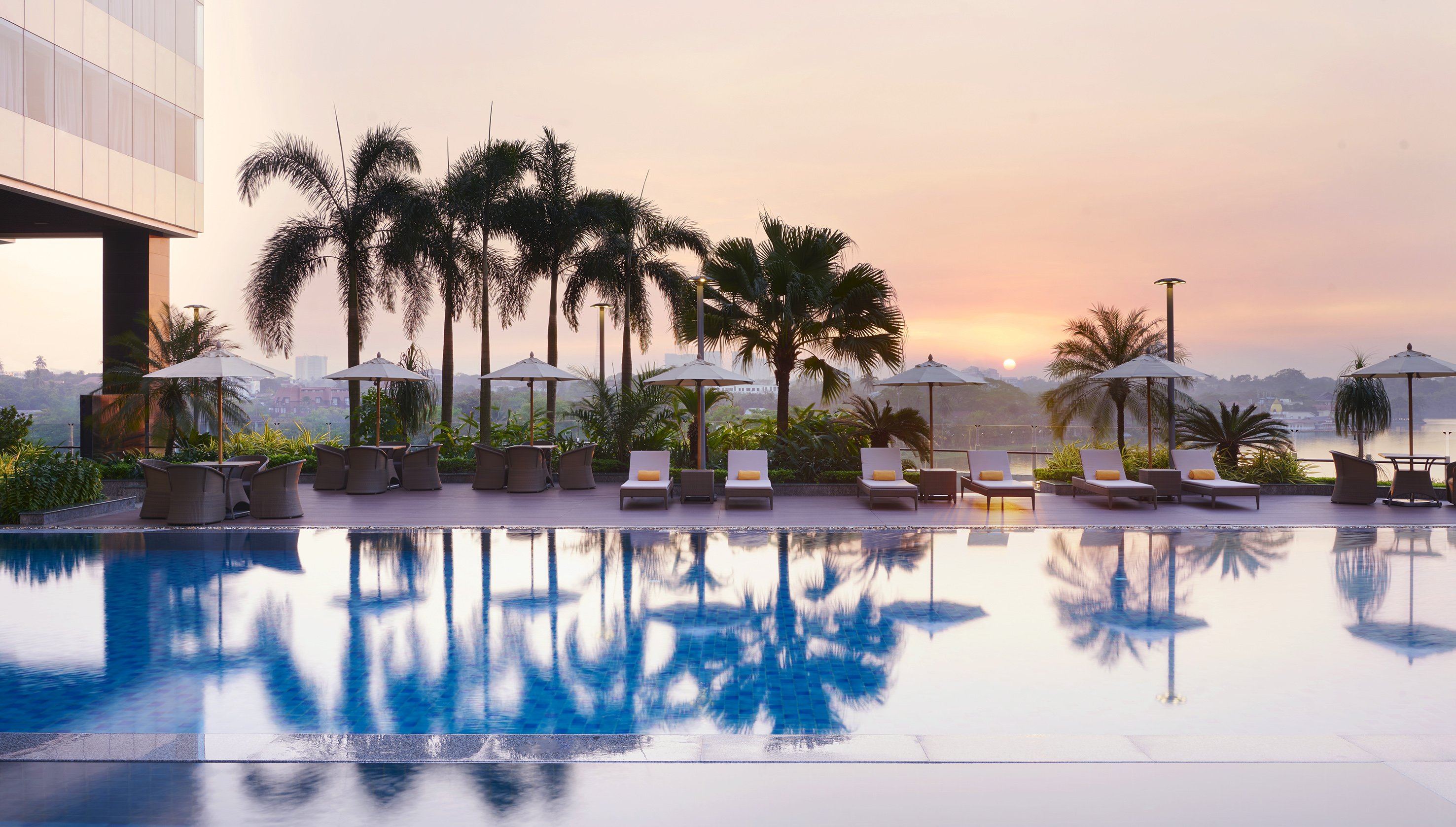 Luxury resort pool with palm trees, lounge chairs, and sunset over water.