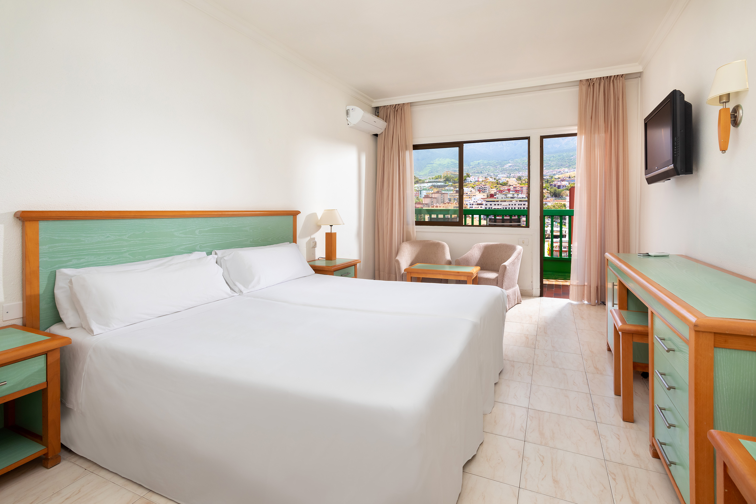Bright hotel room with twin beds, mountain view balcony, green accents.