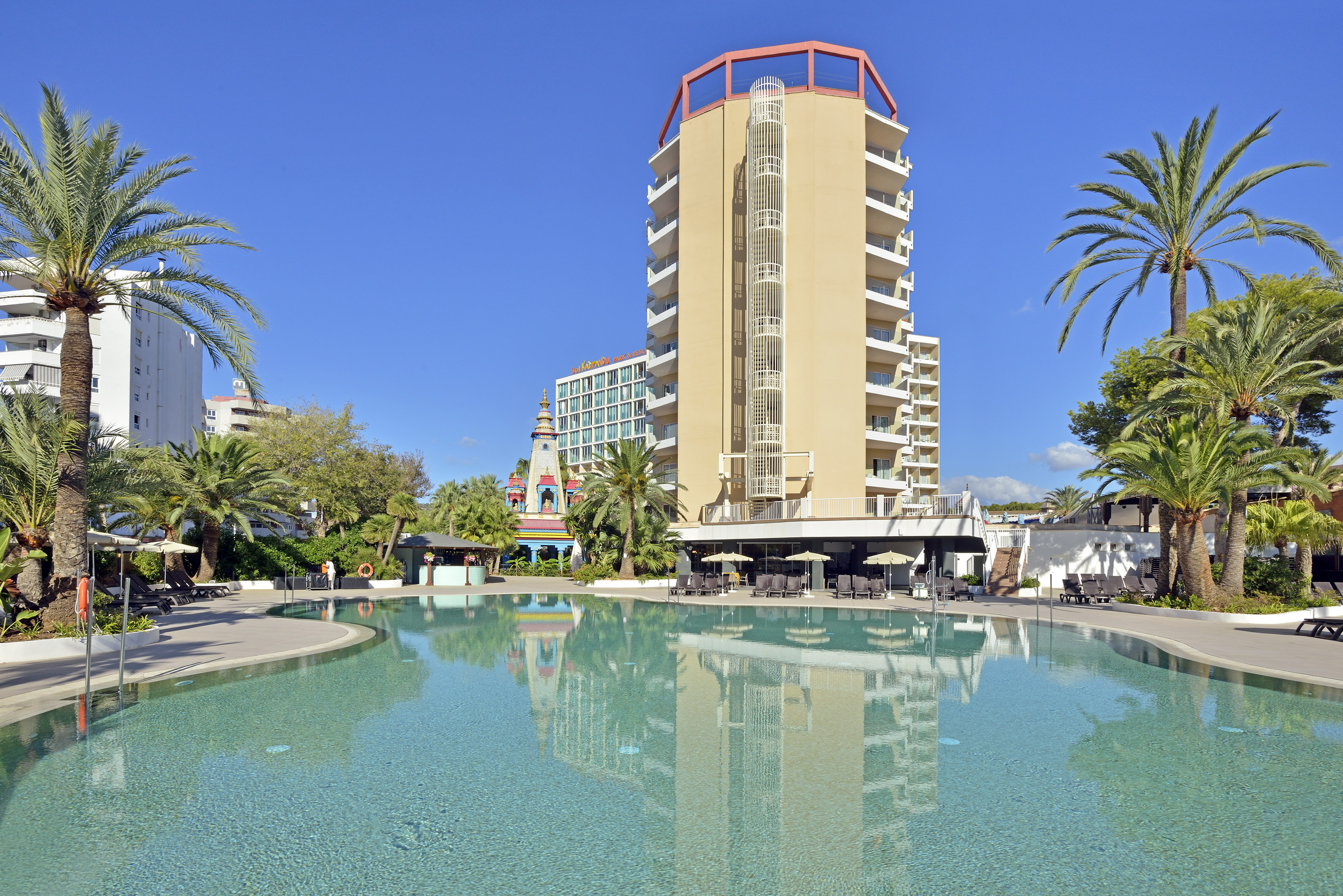 Hotel resort swimming pool with palm trees, modern building, and sunny sky.