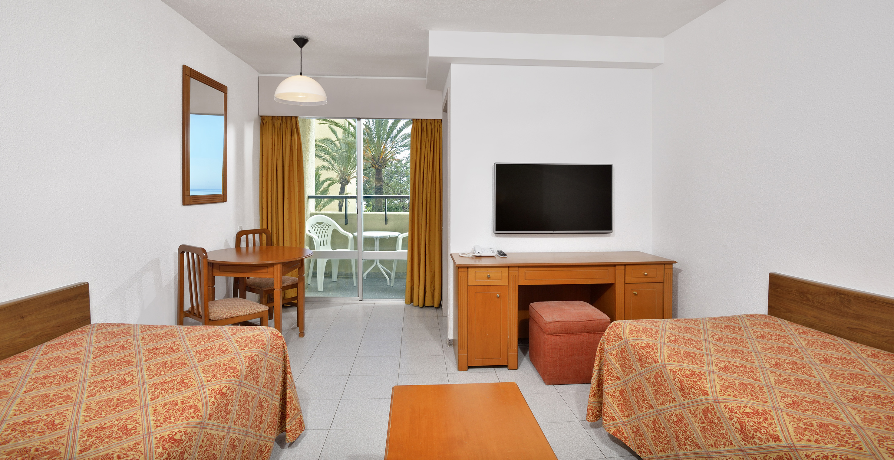 Hotel room with two single beds, balcony, desk, TV, and tropical palm view.