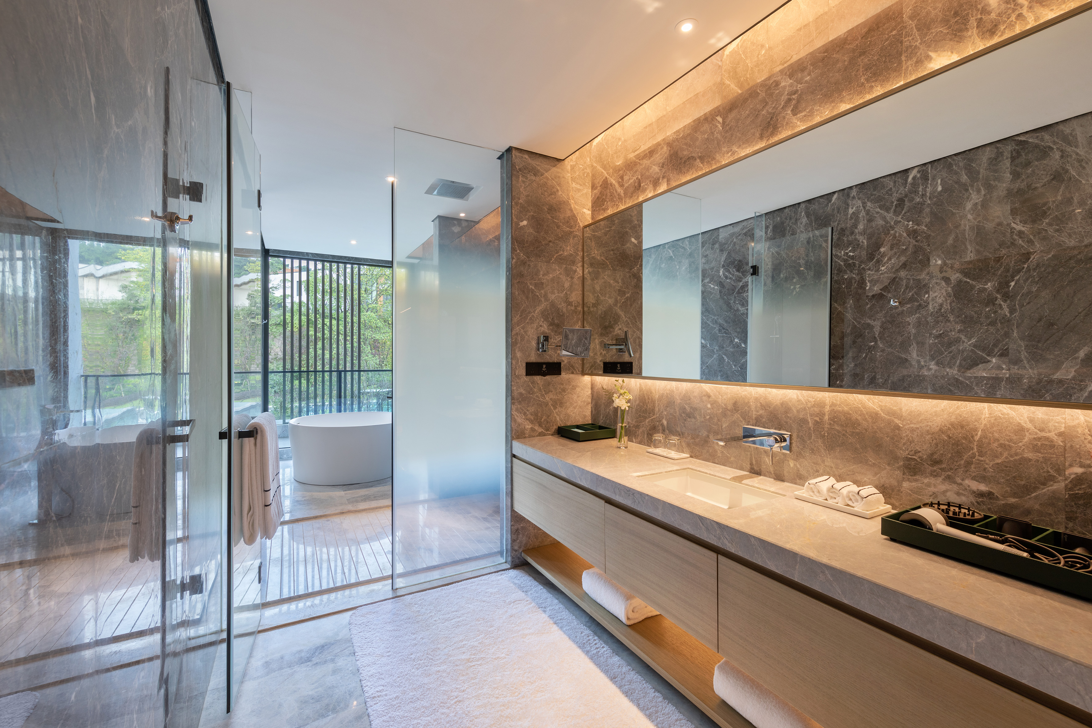 Modern marble bathroom, glass shower, freestanding tub, illuminated vanity.
