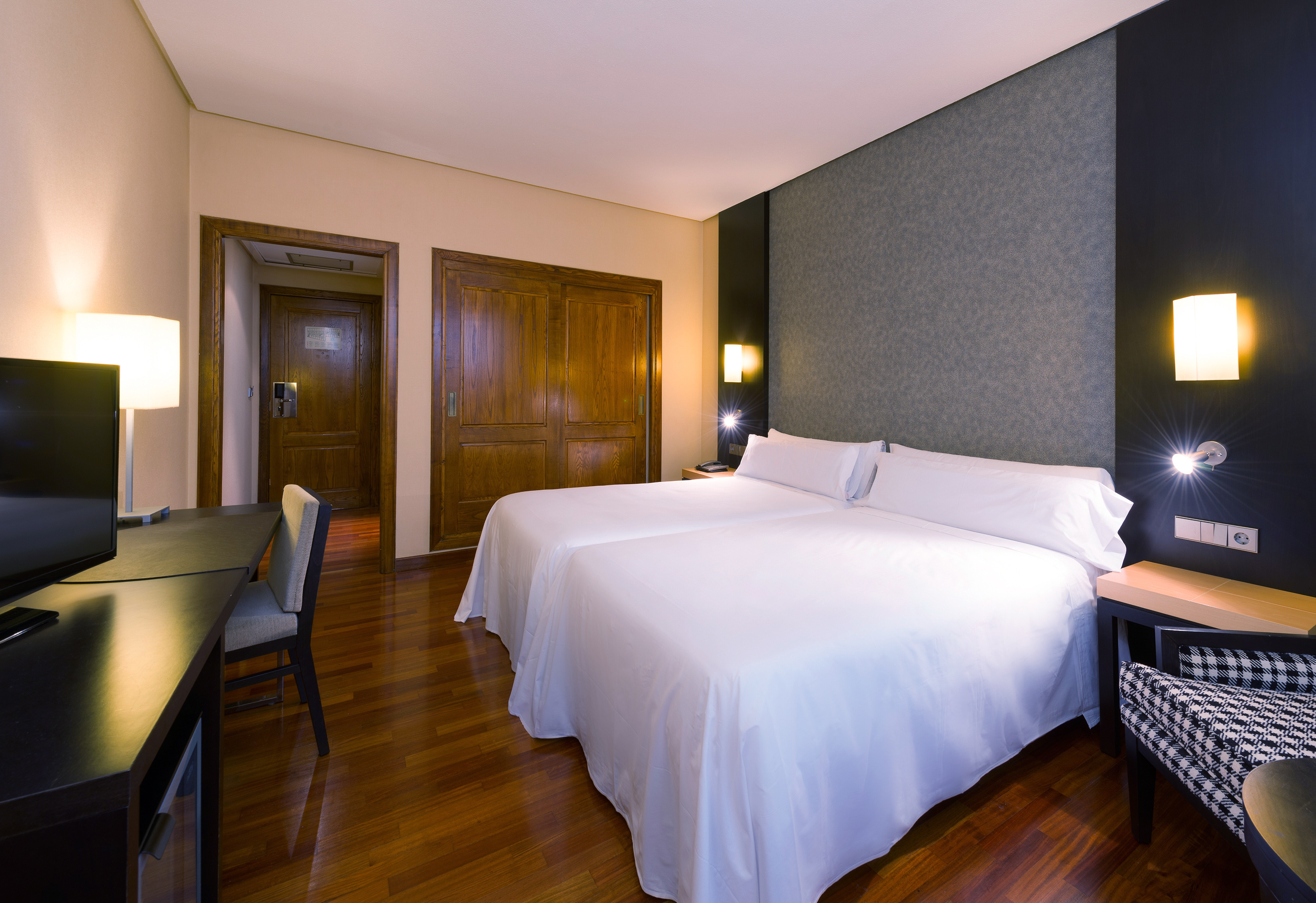 Hotel room with two white beds, dark accent wall, wood floor, desk, and wardrobe.
