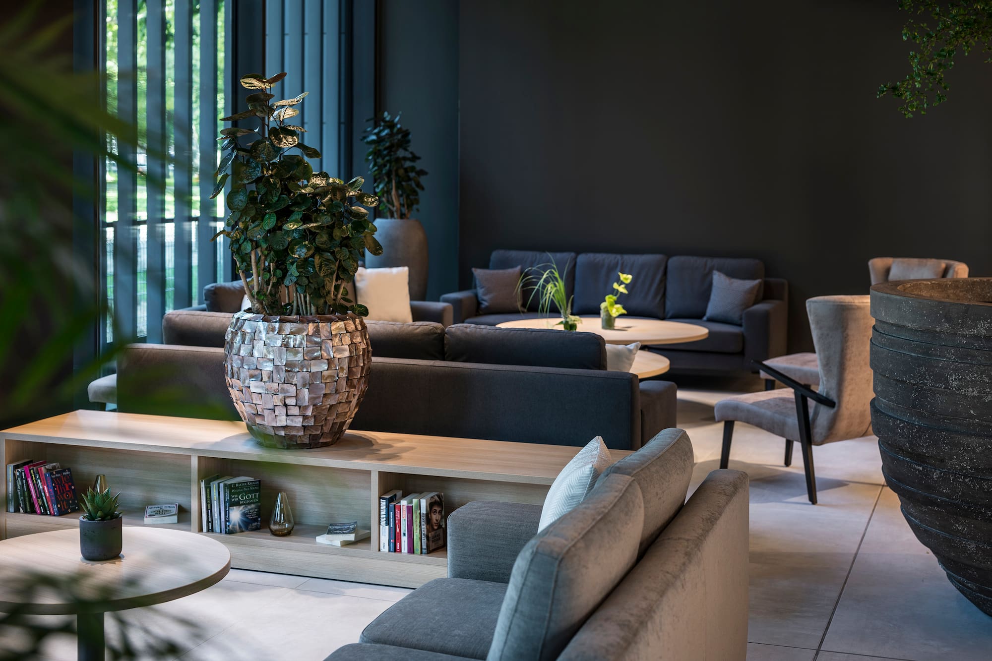 Modern dark lounge with gray sofas, wooden tables, and green plants.