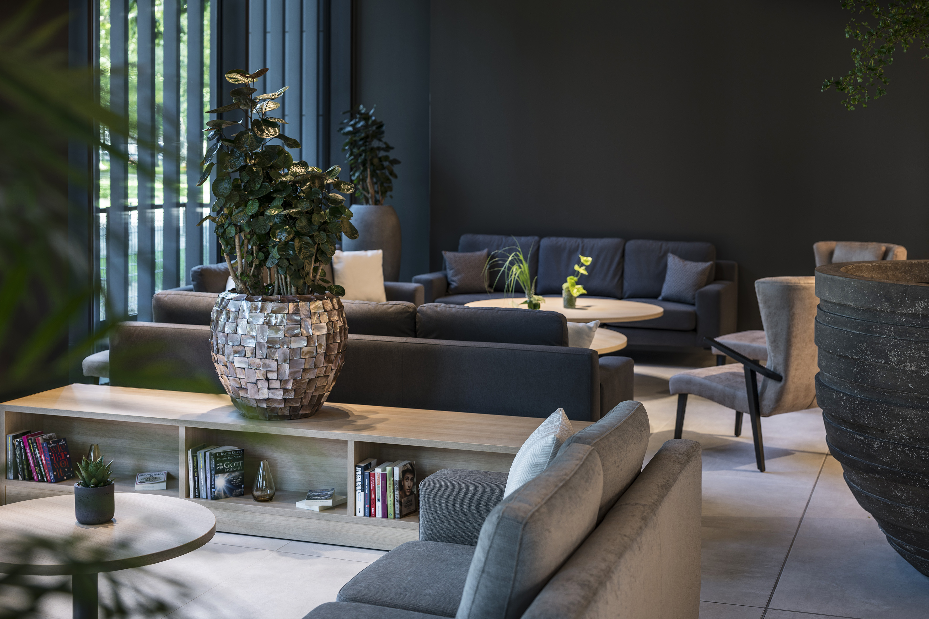 Modern dark lounge with gray sofas, wooden tables, and green plants.