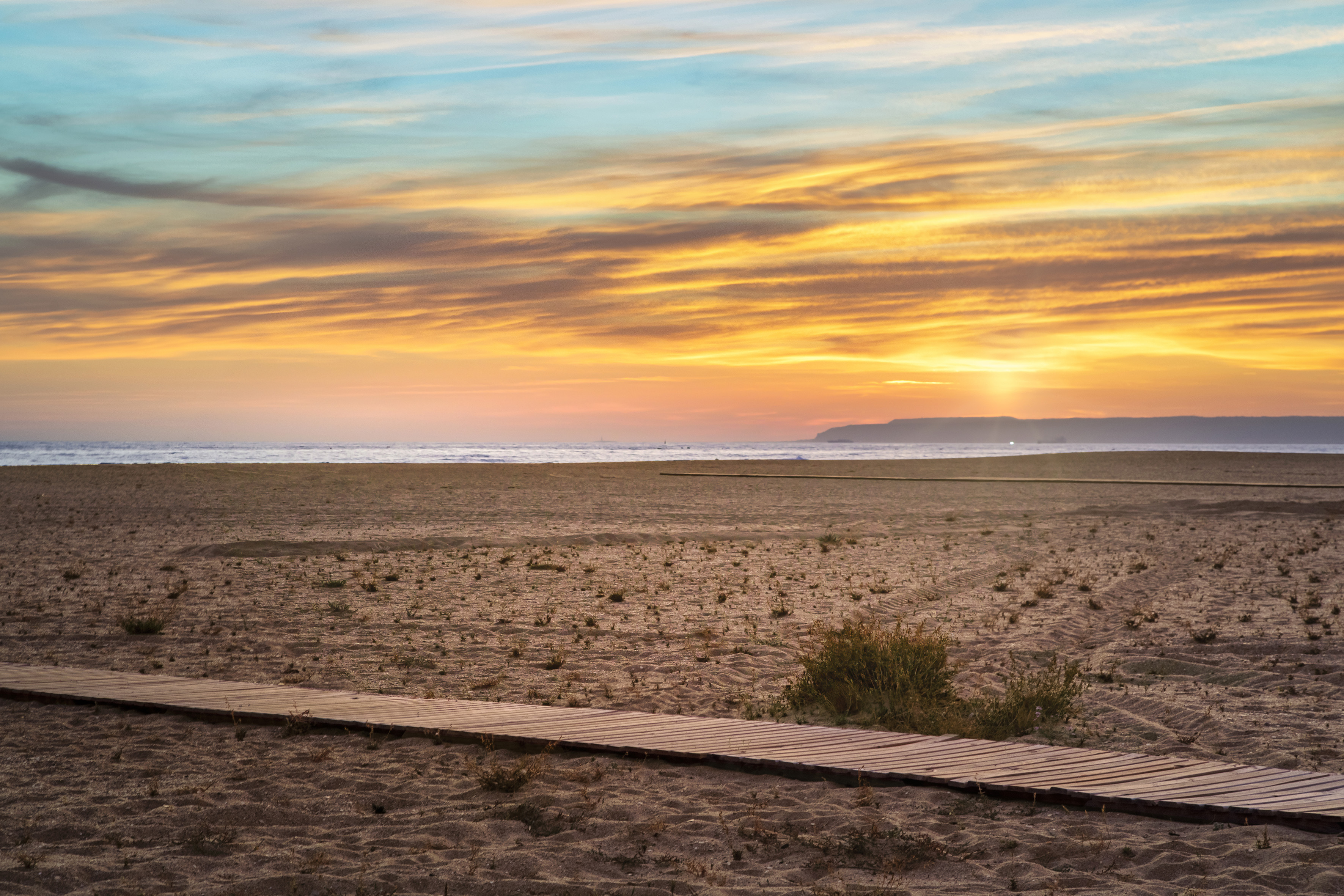 Sunset at sandy beach, ocean, vibrant sky, and wooden boardwalk.