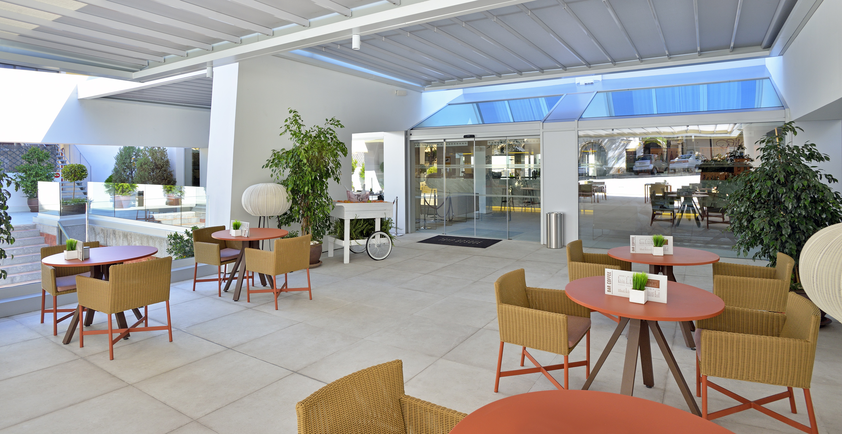 Modern hotel terrace with outdoor tables, wicker chairs, and glass entrance.
