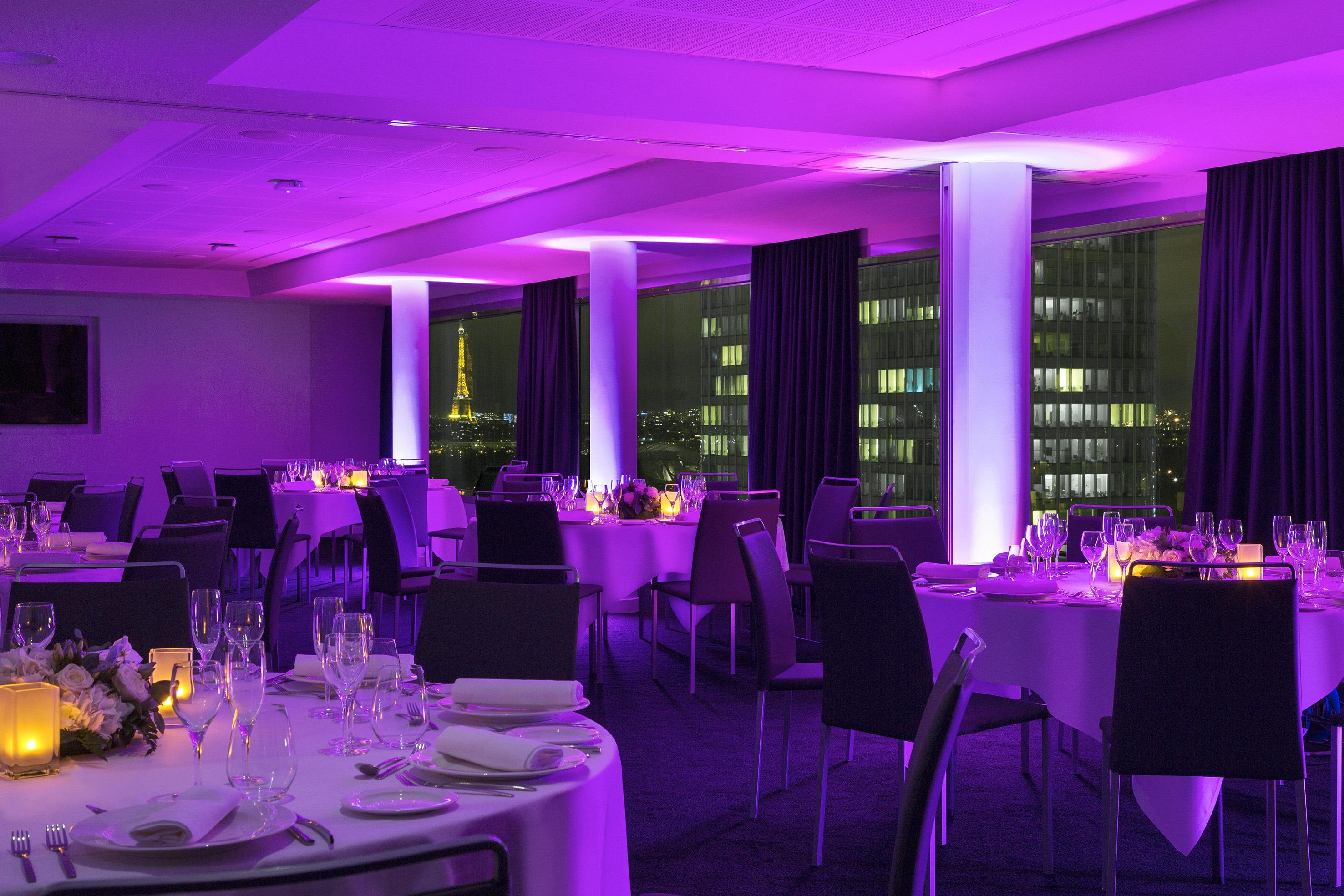Purple-lit event dining room in Paris with set tables and Eiffel Tower view.